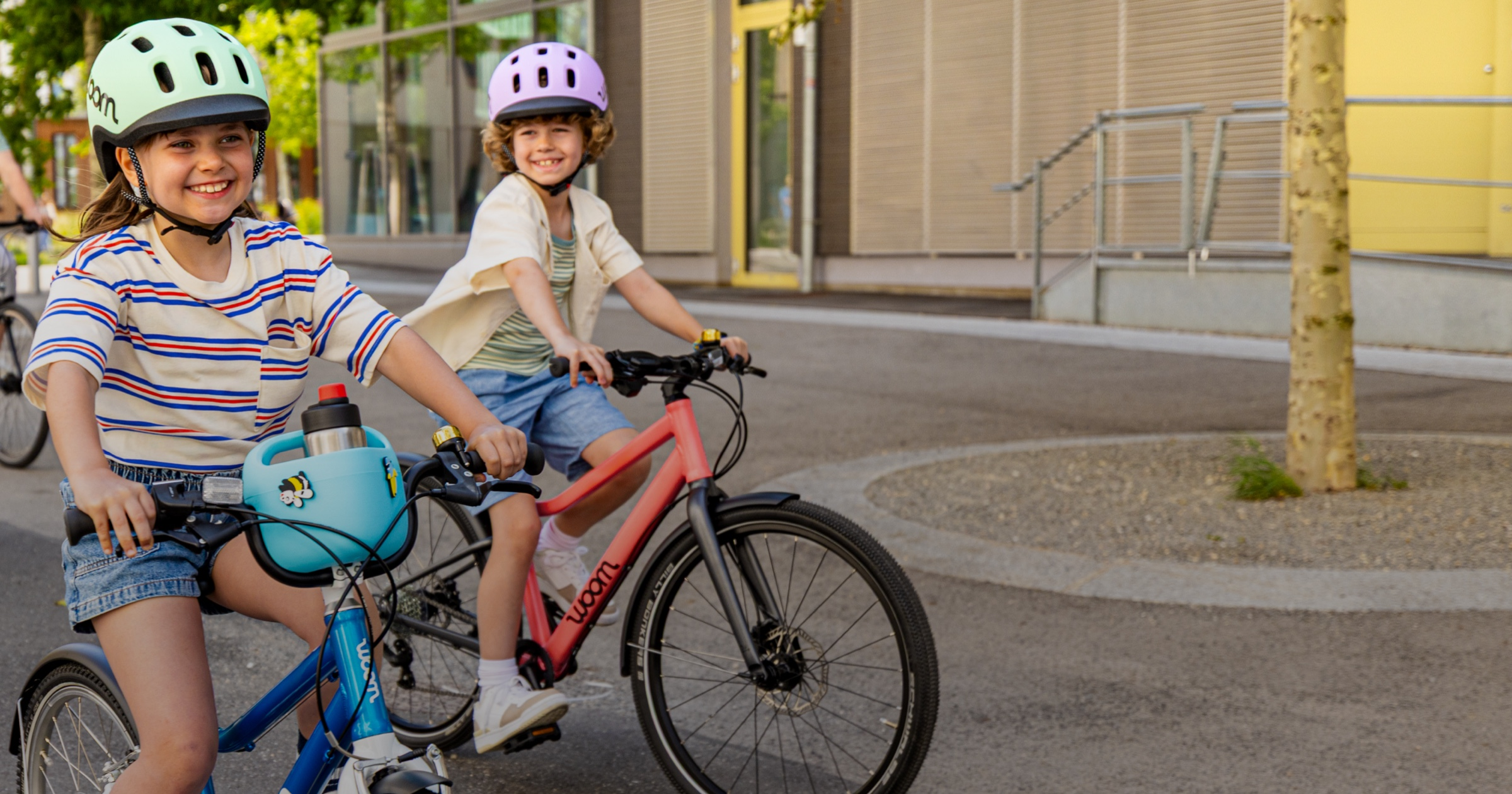Two kids in woom READY Helmets ride their woom bikes, one with a POP Kid’s Bike Basket and GLUG Stainless Steel Water Bottle, down an empty street on a sunny day.