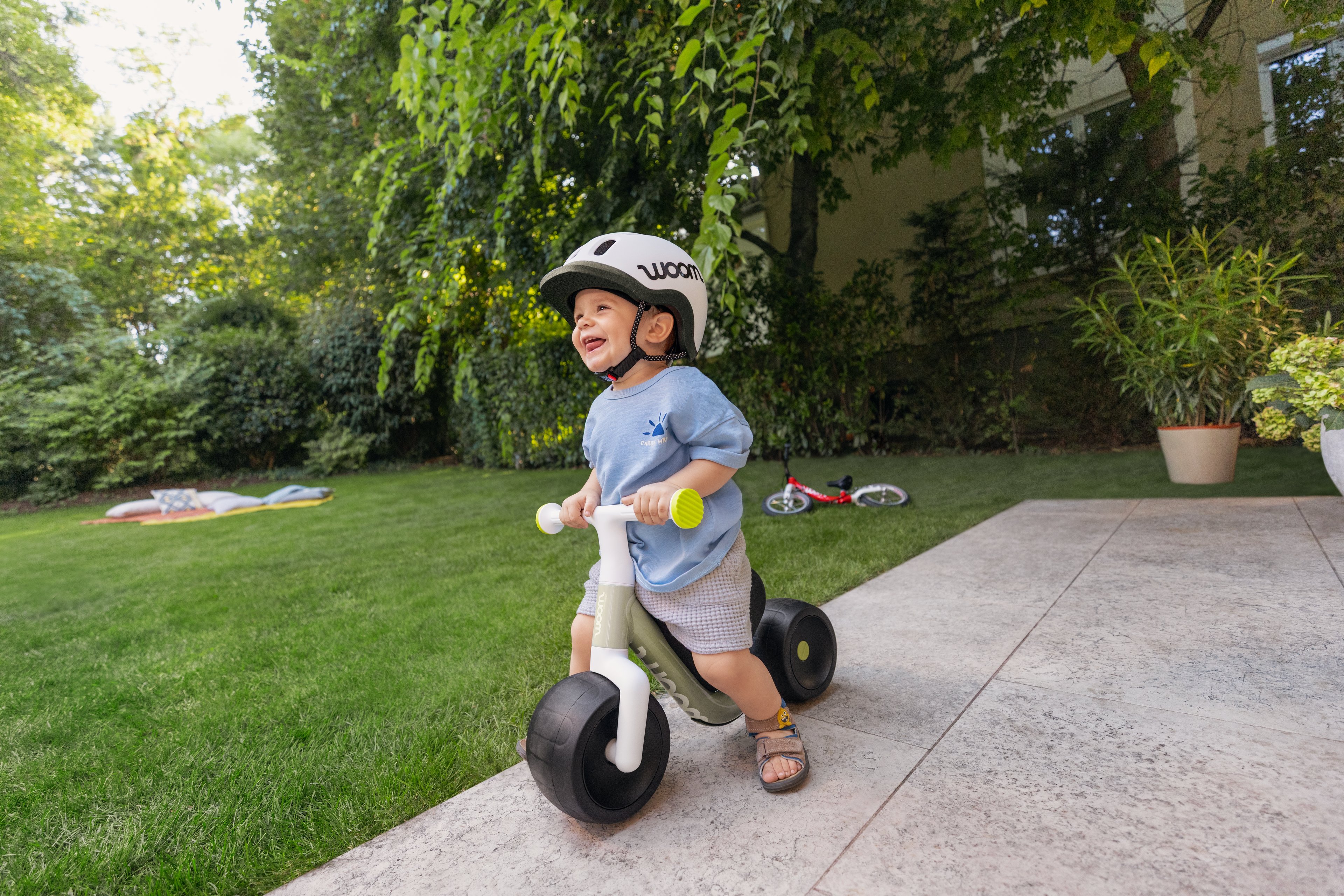 Een lachende peuter met een helm rijdt op een woom WOW loopfiets in de kleur soft sprout op een terras naast een gazon in een groene tuin.