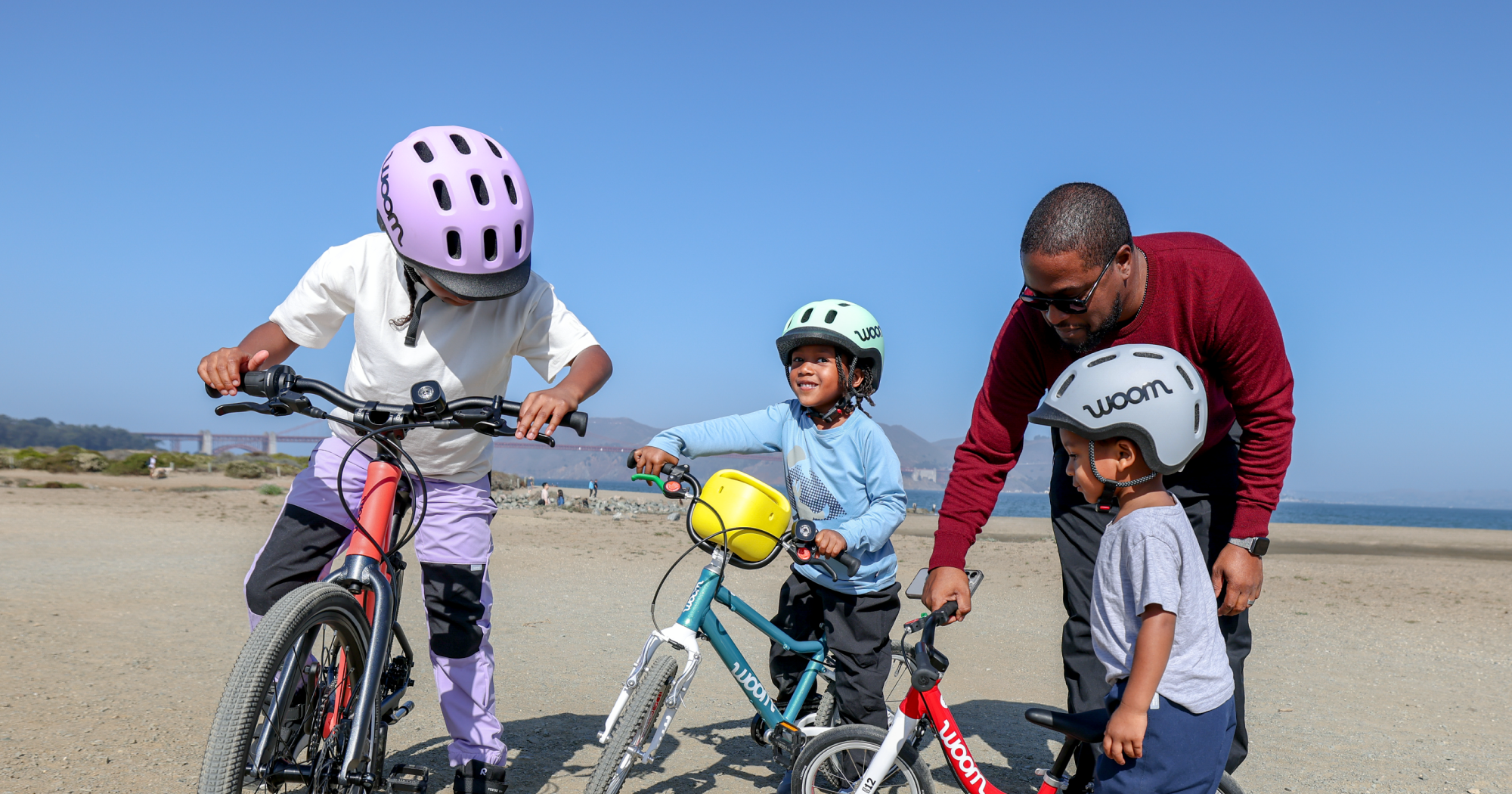 Three kids in READY Helmets prepare to ride their woom bikes on the beach with the help of their dad.