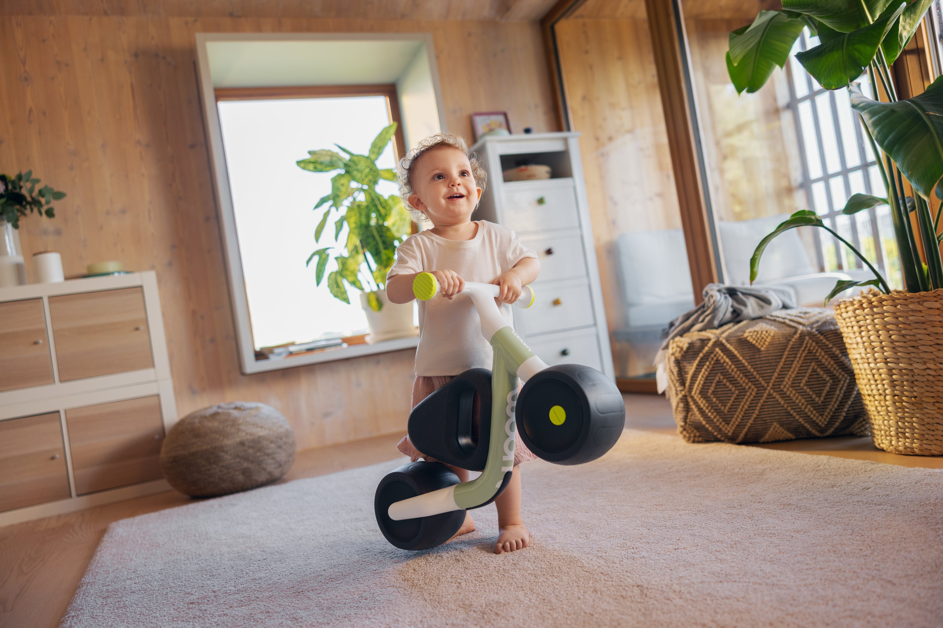 Een blije, blotevoetenpeuter rijdt op een woom WOW loopfiets in soft sprout over een vloerkleed in een lichte, zonovergoten kamer met grote ramen.