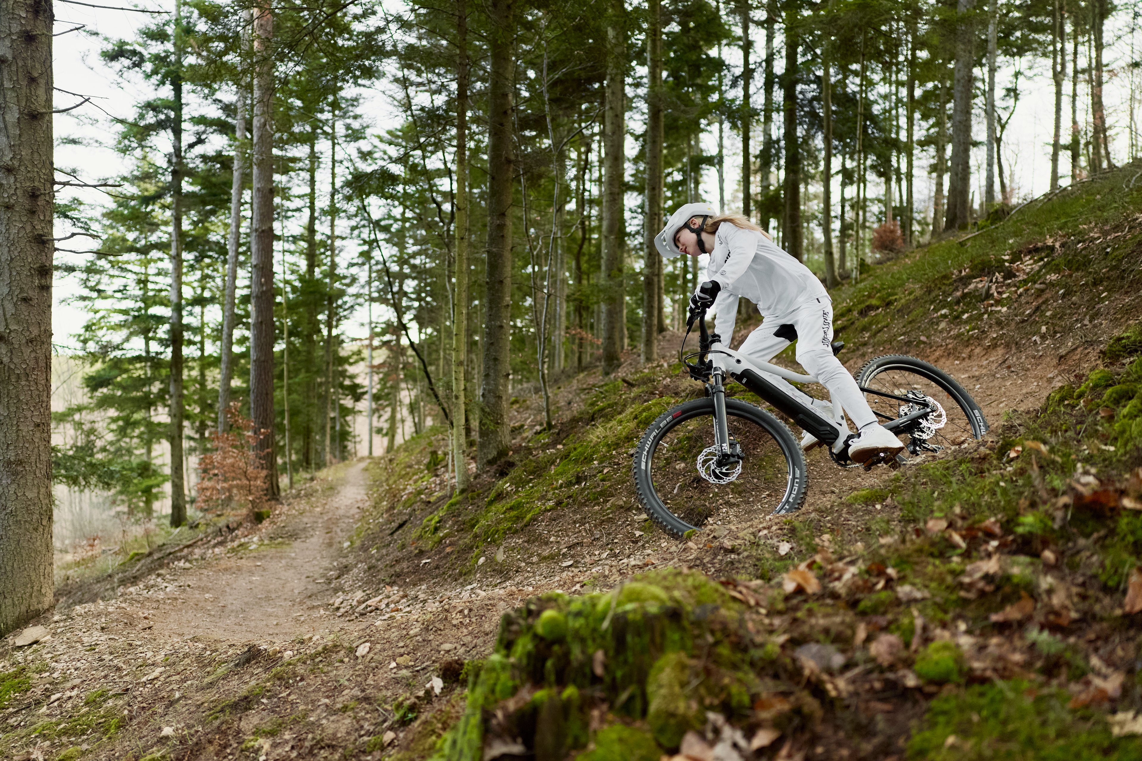 Ein in Weiß gekleidetes Mädchen mit Helm auf dem Kopf fährt mit einem woom UP E-Mountainbike rasant in eine Kurve eines bewaldeten Trails.