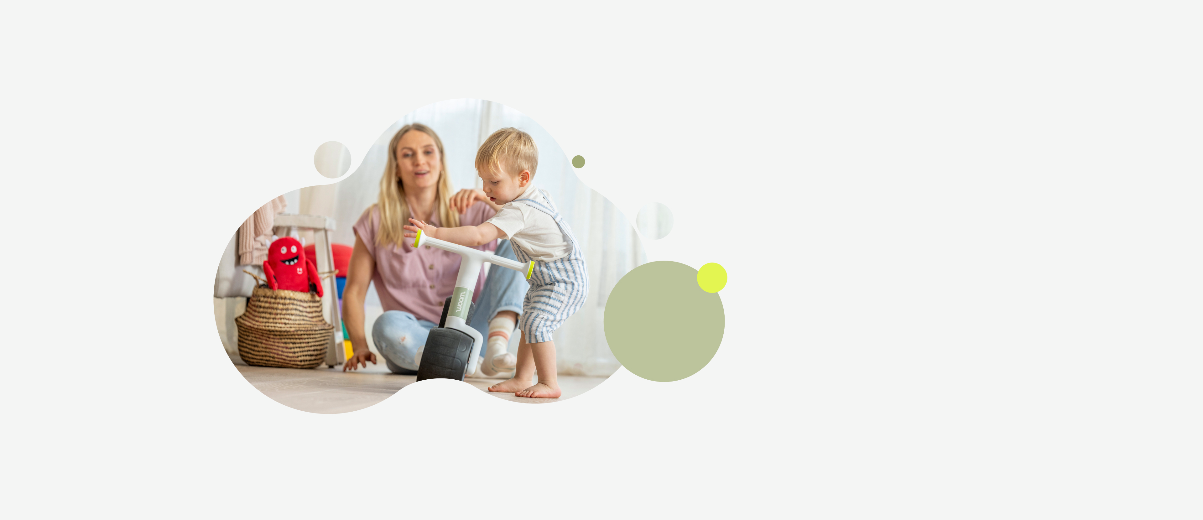 A person stands with a green woom WOW and white ride-on toy indoors, another person sits in the background.