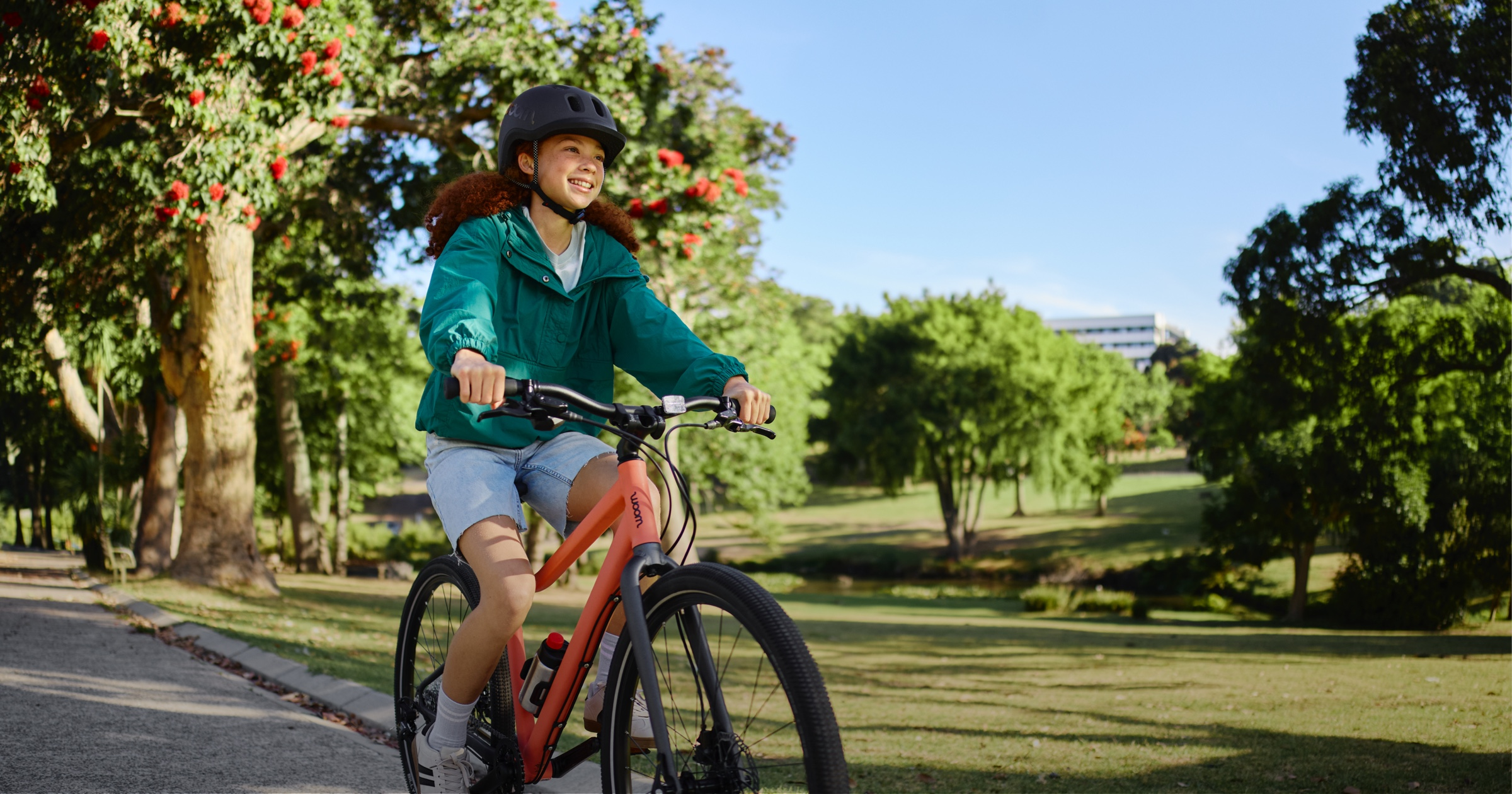 Girl in a black READY helmet with curly red hair, teal windbreaker, and denim shorts rides a neon coral woom EXPLORE bike into the left side of the image. She is in a vibrant, tree-lined park on a sunny day.