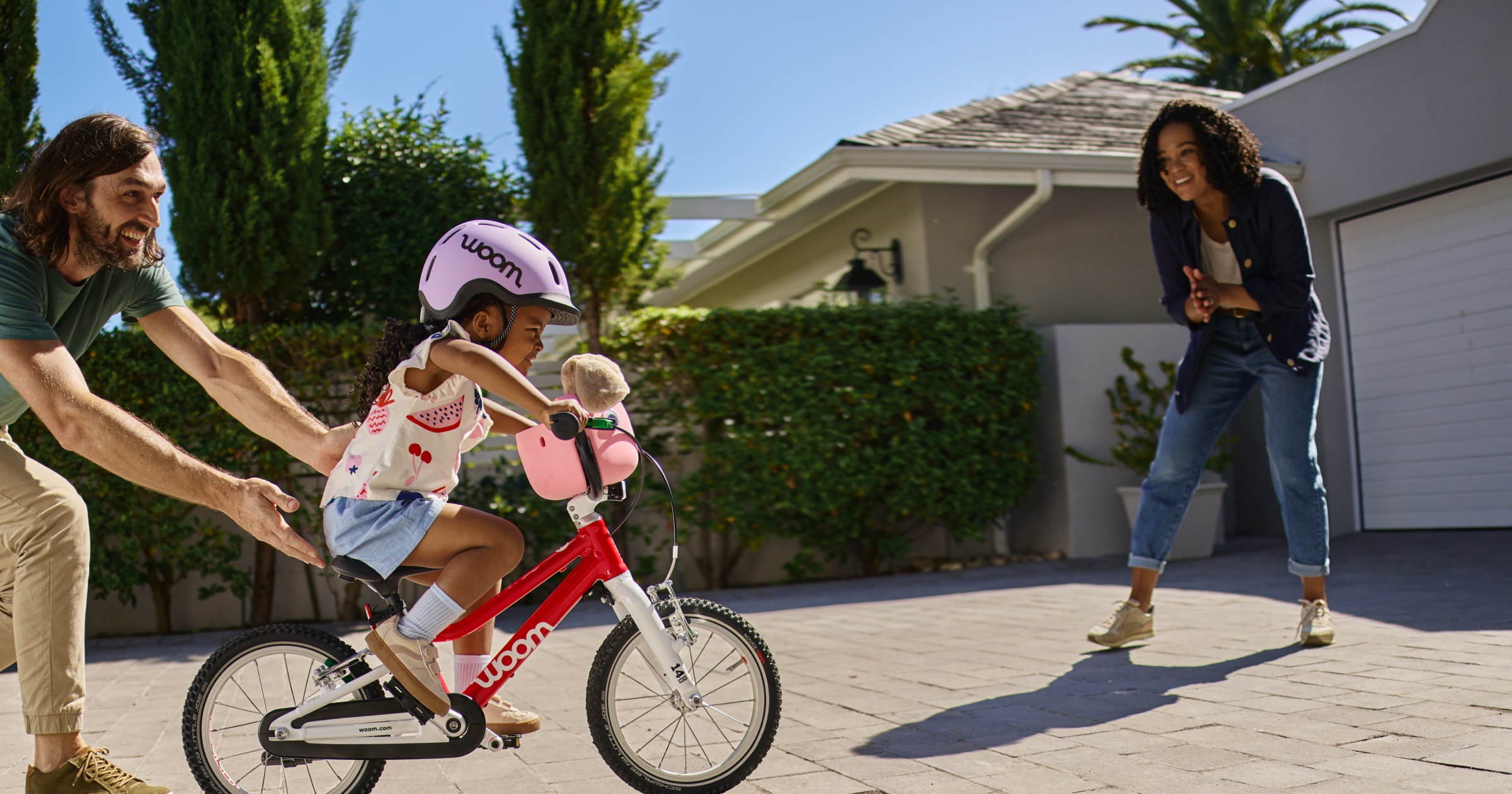 A father guides his young daughter on her woom GO 2 bike toward her mother. They are standing in the driveway and the young girl is wearing a READY Kids’ Helmet.
