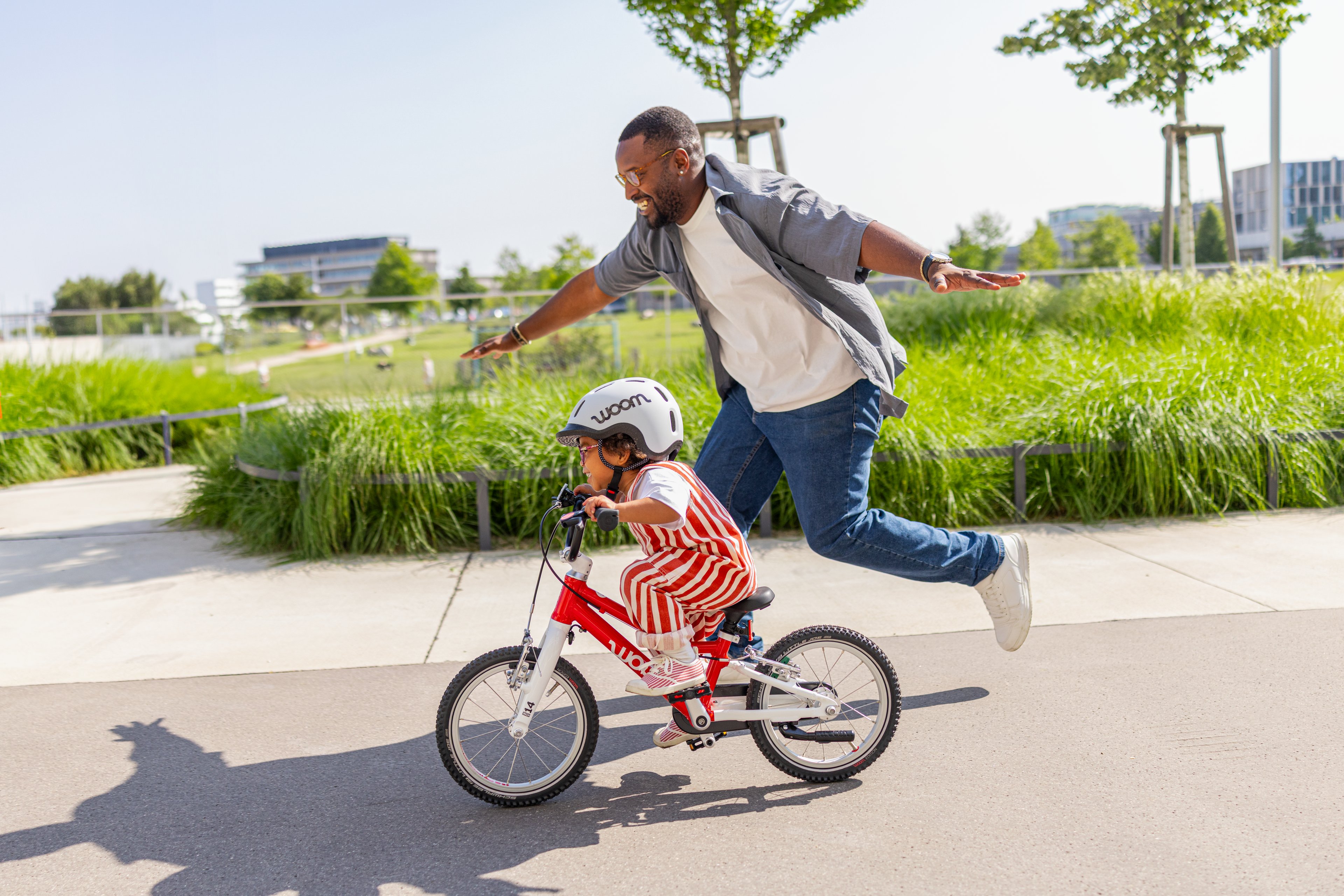 A father runs cheerfully next to his young son as he successfully pedals his red woom GO 2 down an empty street next to a park.