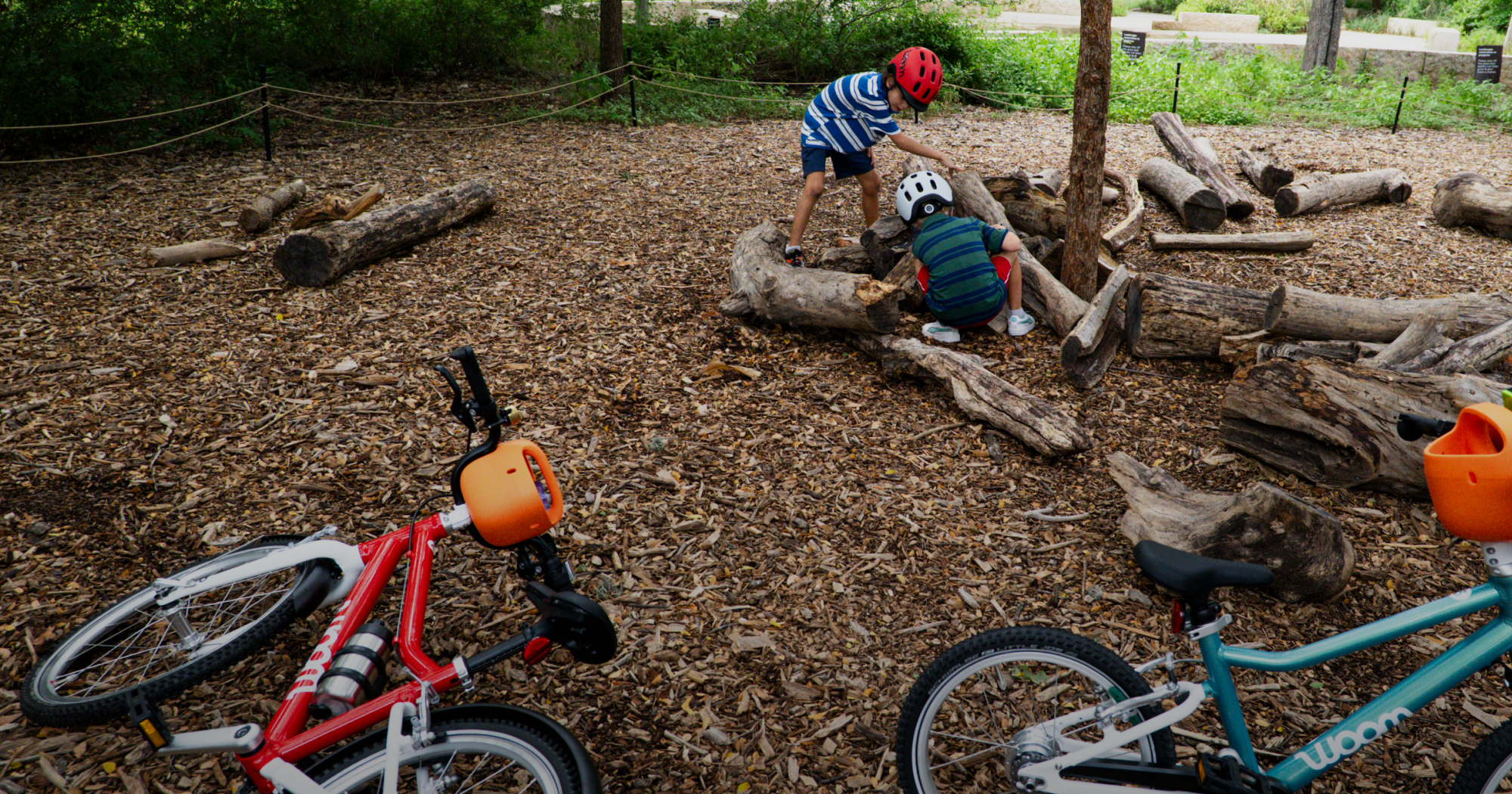 Two boys explore logs in a wooded park area while wearing their woom READY Helmets, with their woom GO bikes laying on the ground nearby.