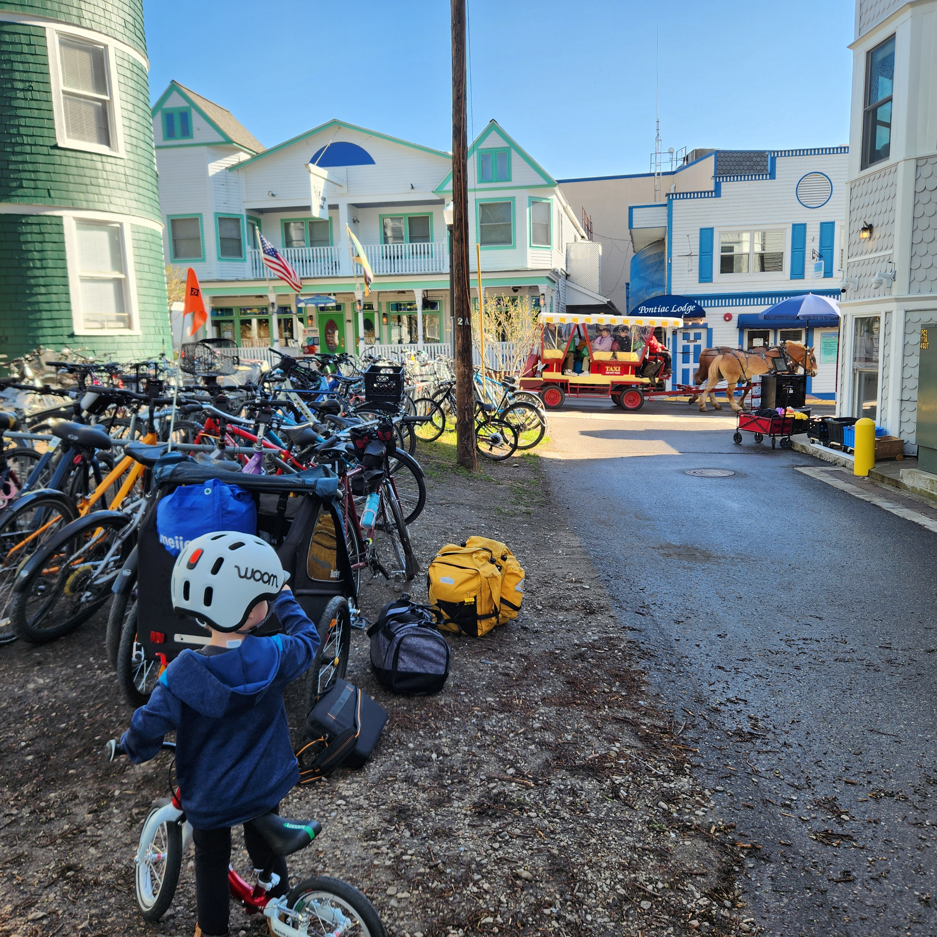 Small boy wearing a light grey READY Kids’ Helmet with READY Helmet Light attached faces away from the camera while watching a horse-drawn taxi pass by. He is standing near a large quantity of locked, colorful bicycles on the side of a quaint street lined with brightly colored stores.