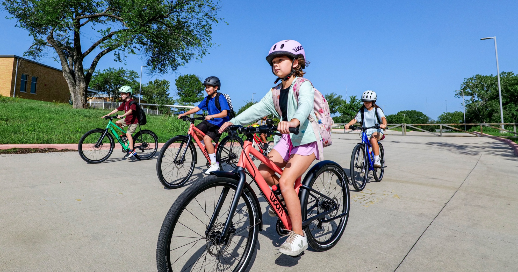 Four kids ride their fully equipped woom bikes through a school parking lot, complete with GLUG water bottles, SNAP click-on fenders, LEGGIE kickstand, and BING bell. Now including BEAM bike lights.