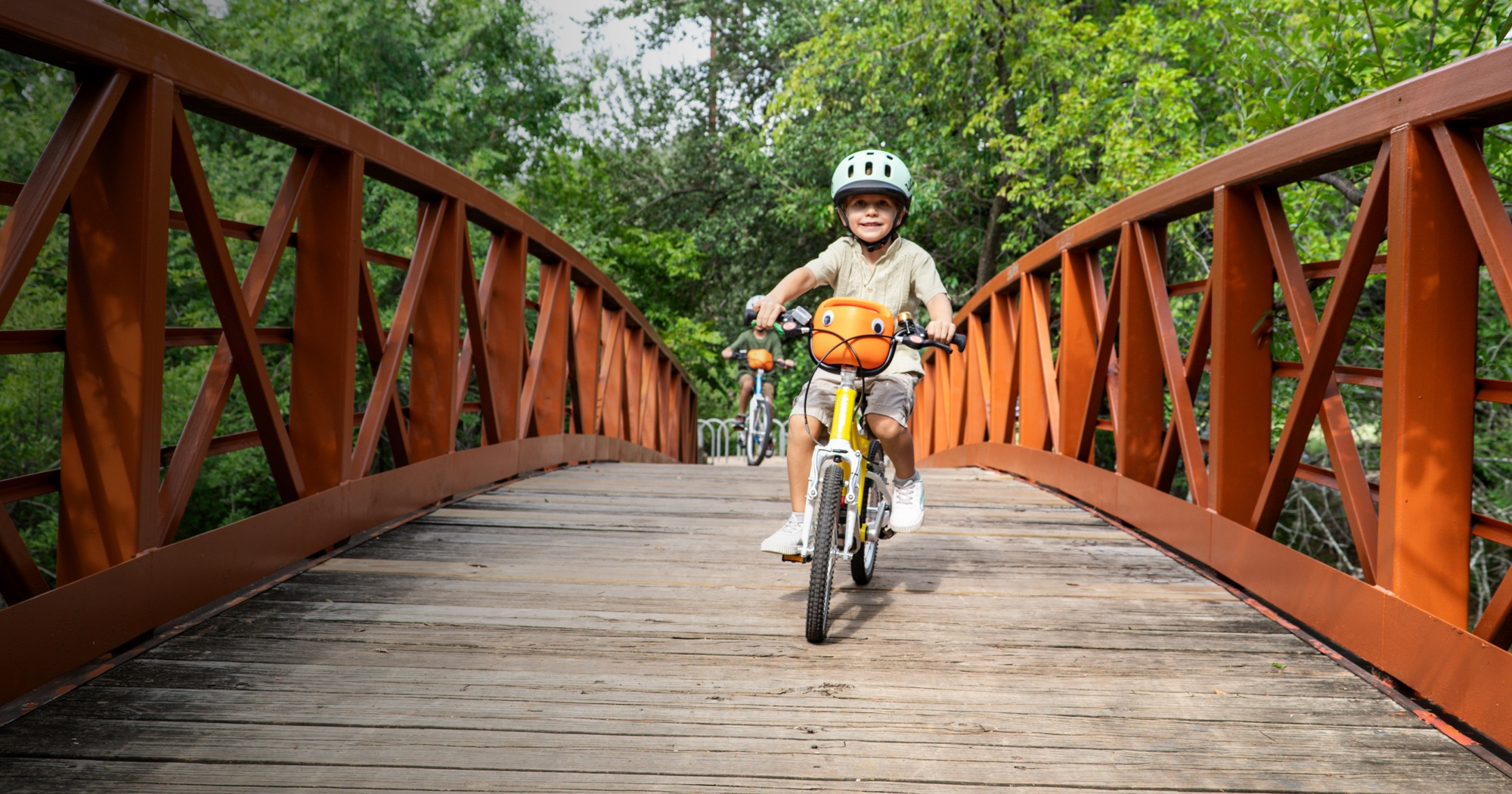 A boy rides a yellow woom GO bike with new orange POP bike basket attached. He is crossing a bridge while wearing a READY helmet, and another young boy rides several paces behind.