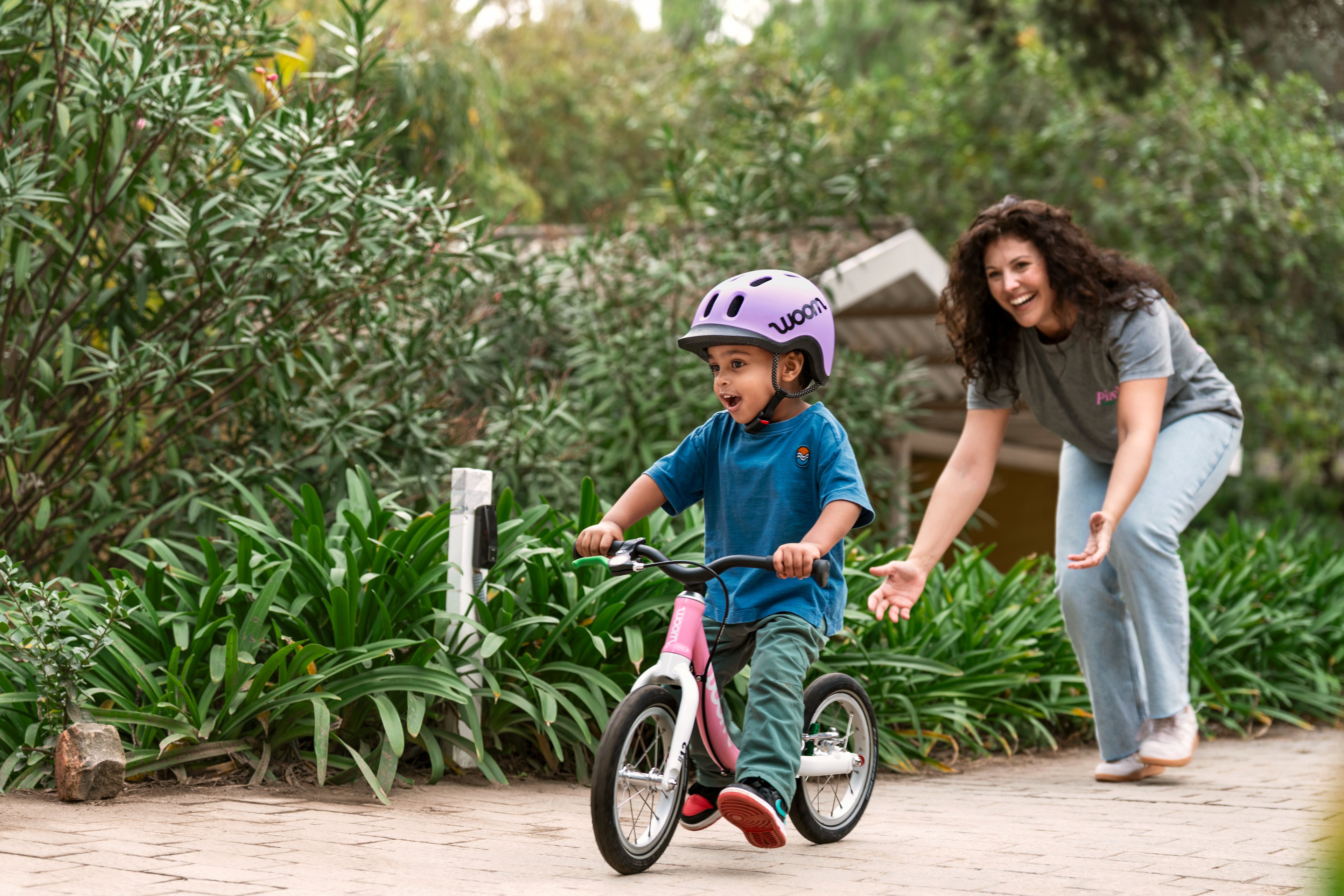 Child in a lilac woom READY helmet on a powder pink woom GO 1 bike, with a woman cheering on a garden path.