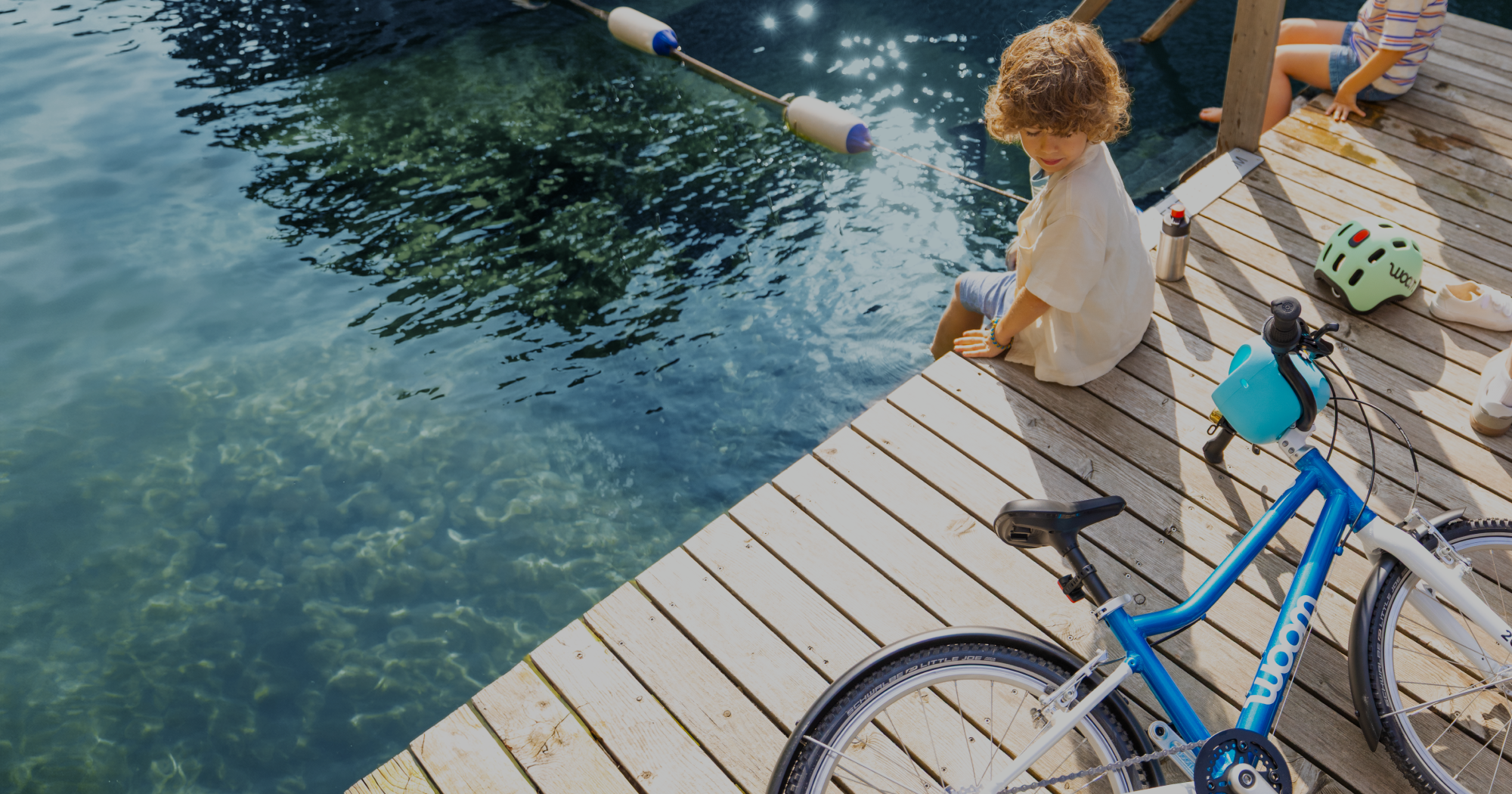 An overhead image of a boy sitting on a dock and dangling his feet into blue water. He’s looking at his metallic blue woom GO 4 lying nearby alongside a GLUG Water Bottle and READY Kid’s Helmet.