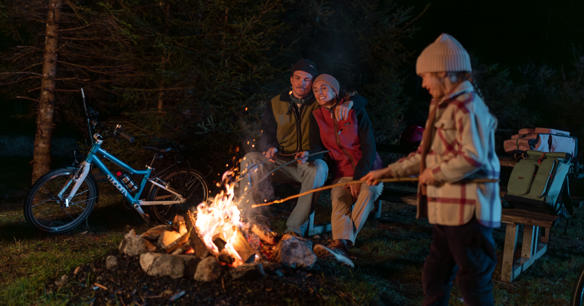 A family gathers around a campfire at night with woom bikes parked at the campsite.
