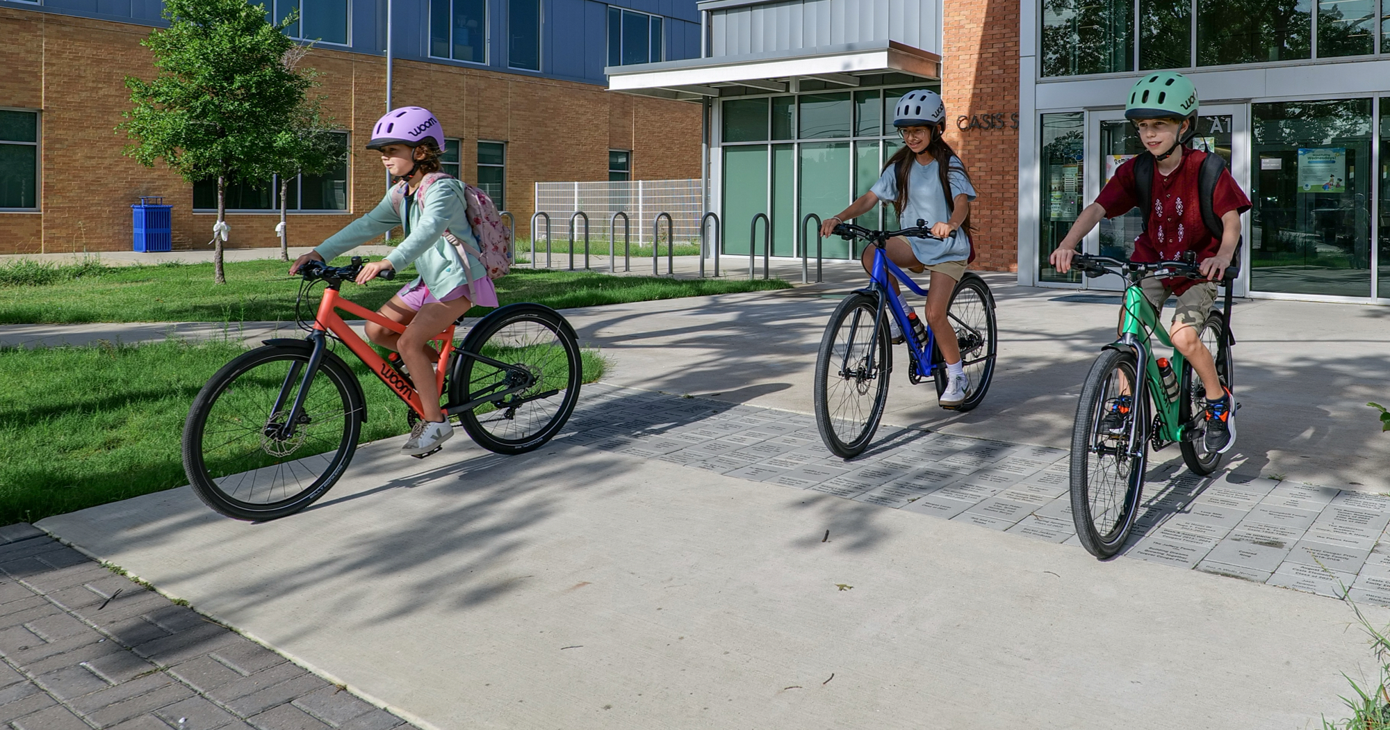 A group of young bike riders steer their woom EXPLORE bikes through a school parking lot. They are wearing woom READY helmets and riding magnetic blue, neon coral, and jungle green woom EXPLORE bikes.