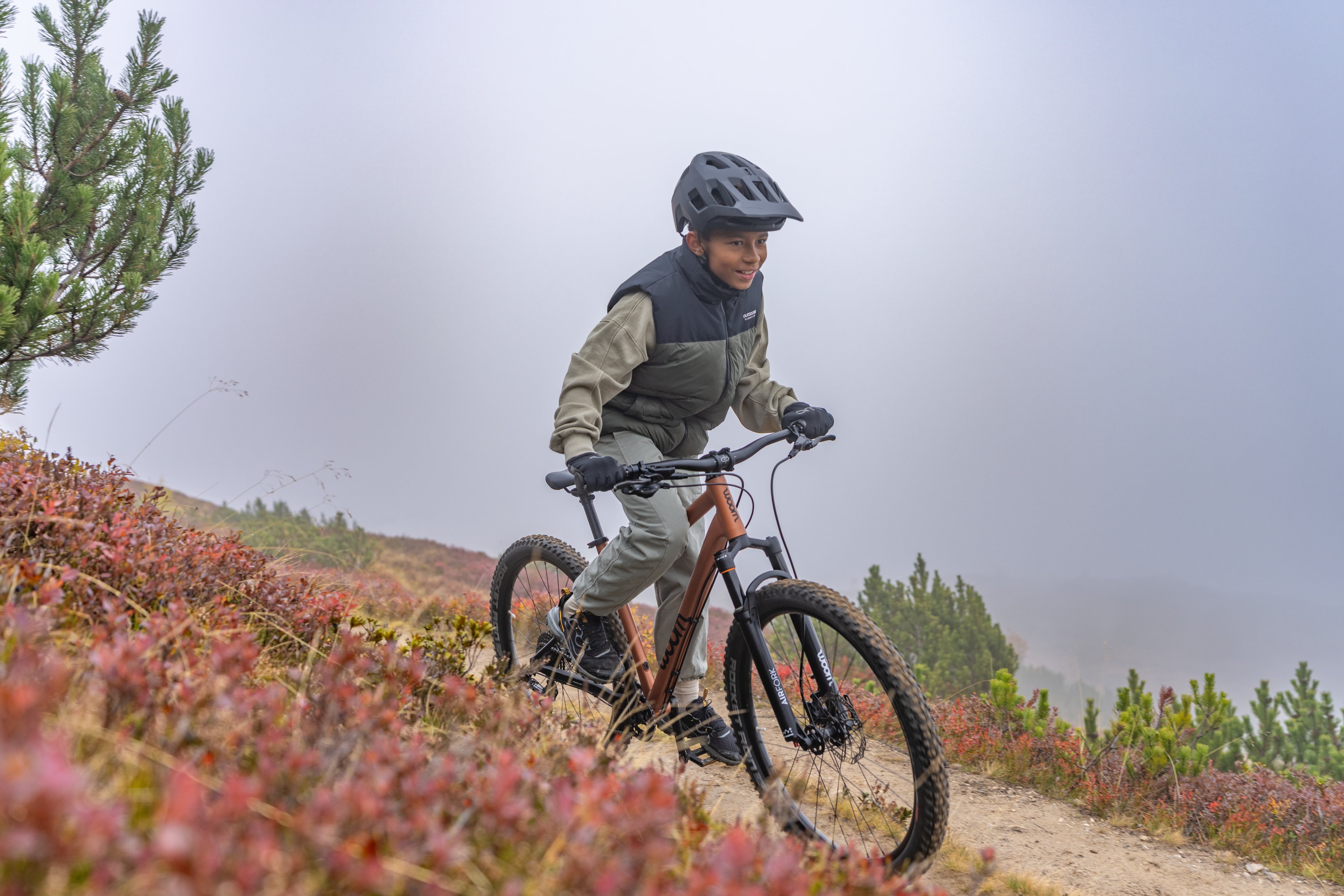 Child wearing a helmet rides a woom OFF mountain bike on a narrow trail through an autumn landscape.
