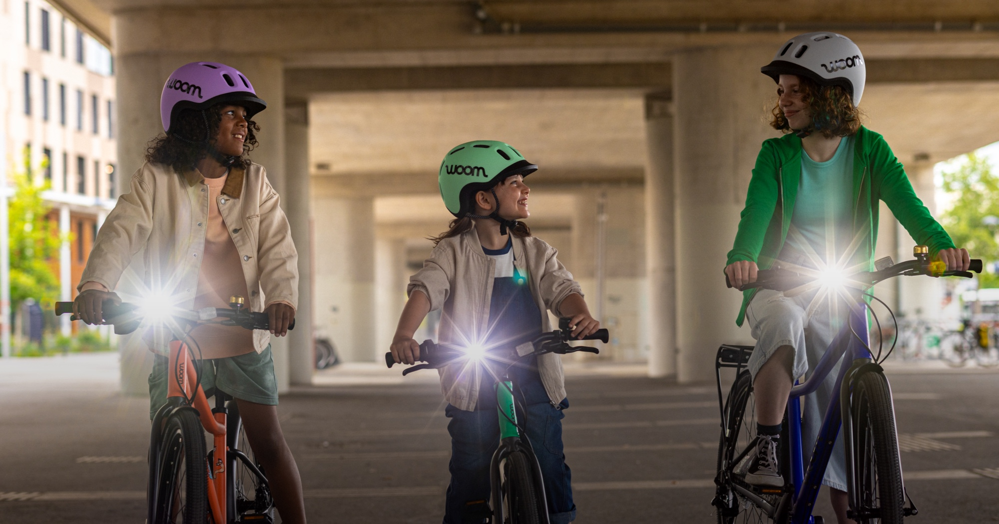 Three kids ride woom bikes in a dimly lit outdoor space with new BEAM Bike Lights attached & shining