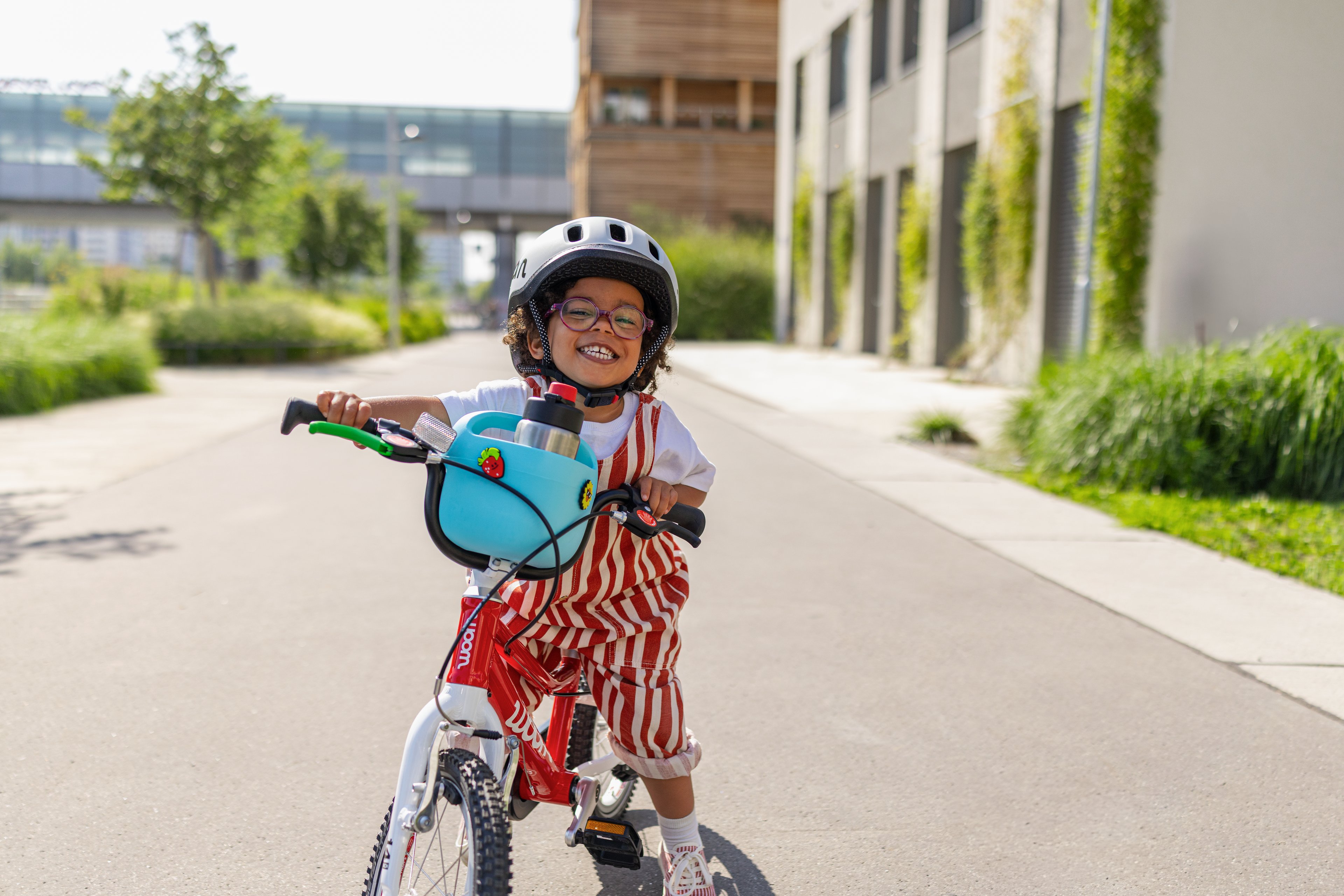 Young boy in a grey READY Helmet rides his red woom ORIGINAL bike, complete with a blue POP Bike Basket and summer POP Ons.