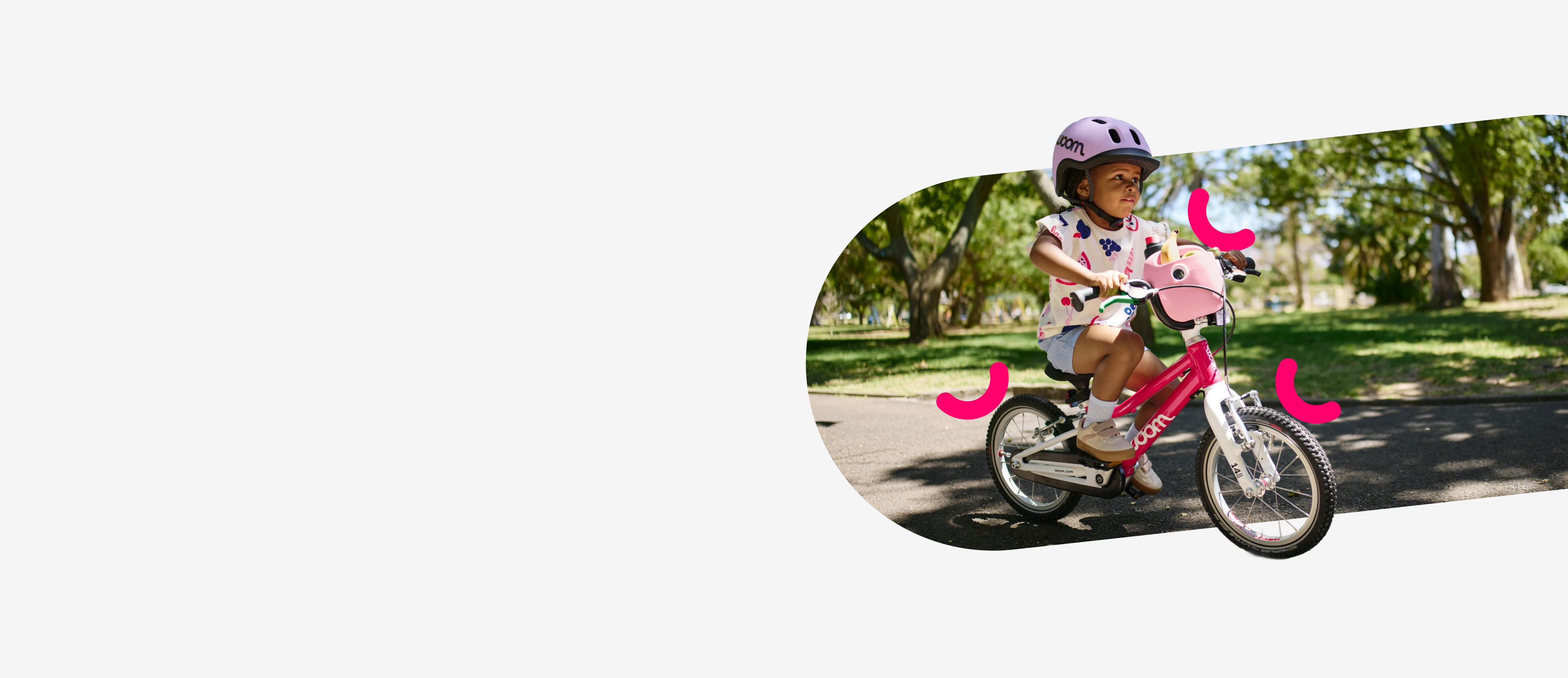 A person wearing a purple READY helmet rides a pink woom GO bicycle with a front POP basket through a park.