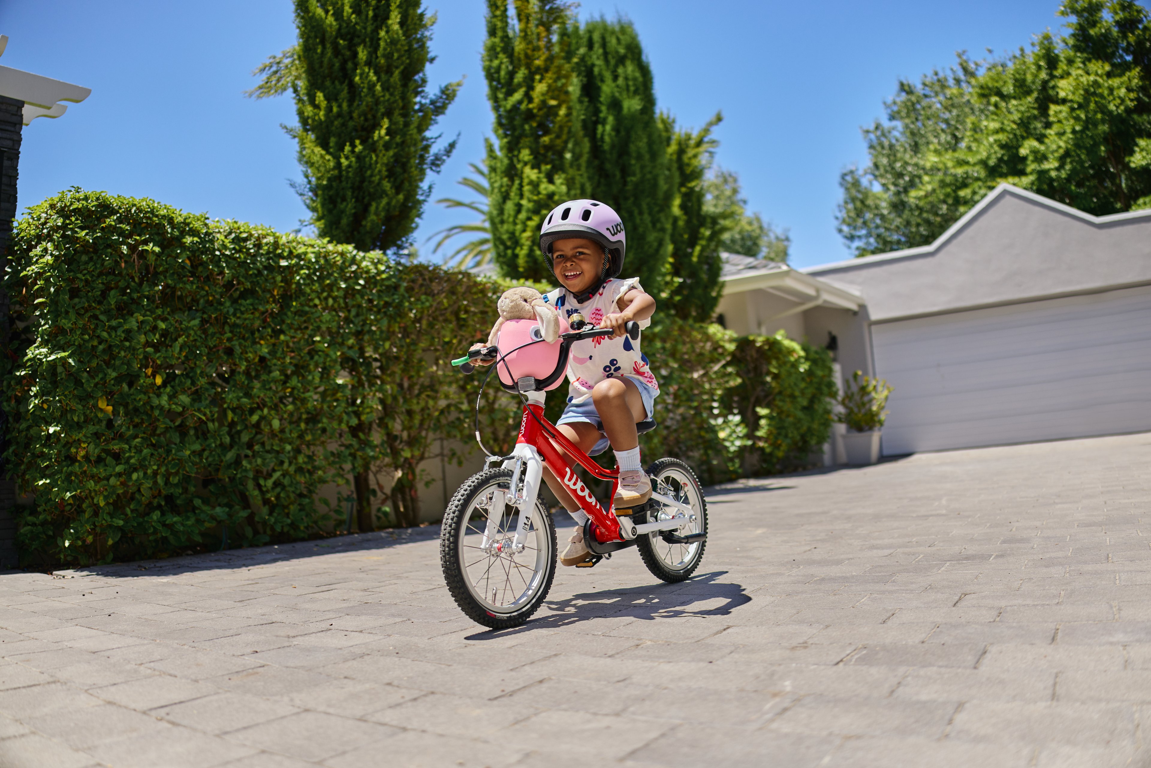 Ein Kind mit lila READY Fahrradhelm auf dem Kopf fährt auf einem roten woom GO 2 Fahrrad in der Hauseinfahrt.