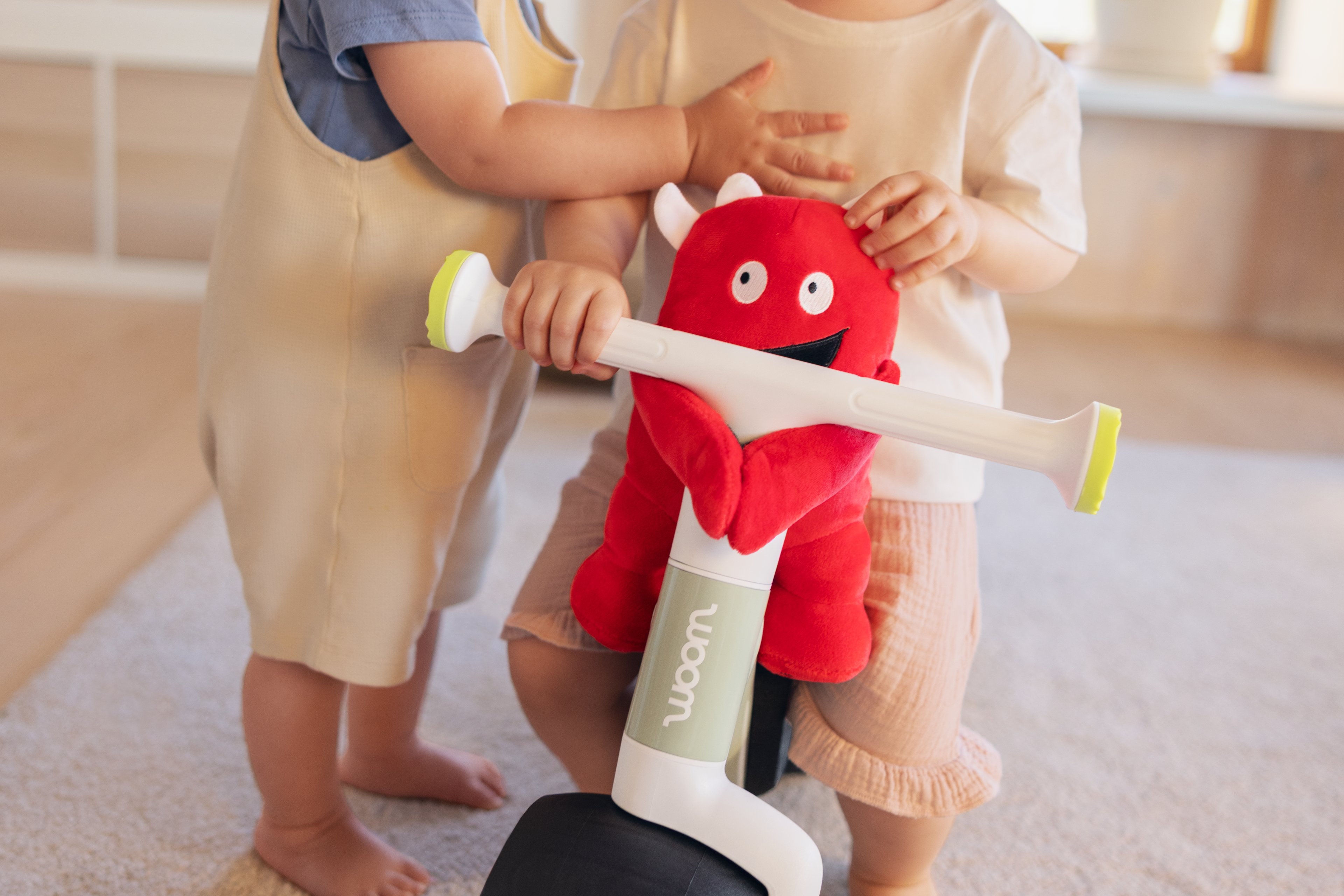 Twee peuters staan bij een woom WOW loopfiets in soft sprout, die versierd is met een rood pluche monster op het stuur.