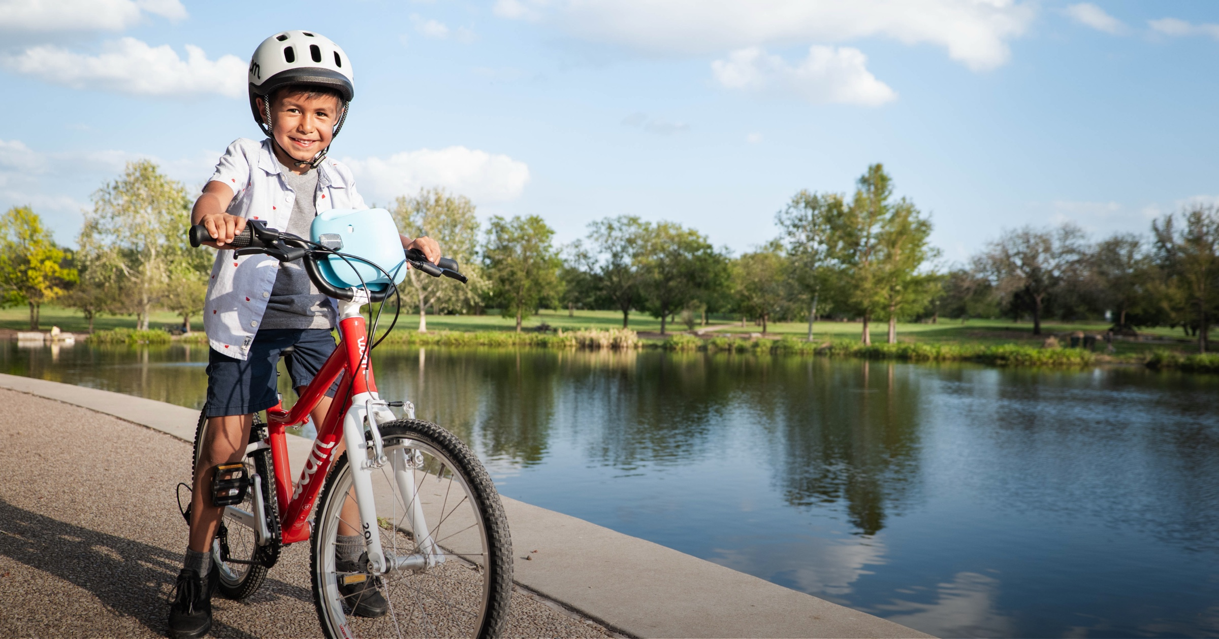 A boy on a paved path next to a tree-lined lake wearing a pale grey READY Kids’ Bike Helmet and riding a red woom GO bike with a bubblegum blue POP Kids’ Bike Basket attached to the handlebars