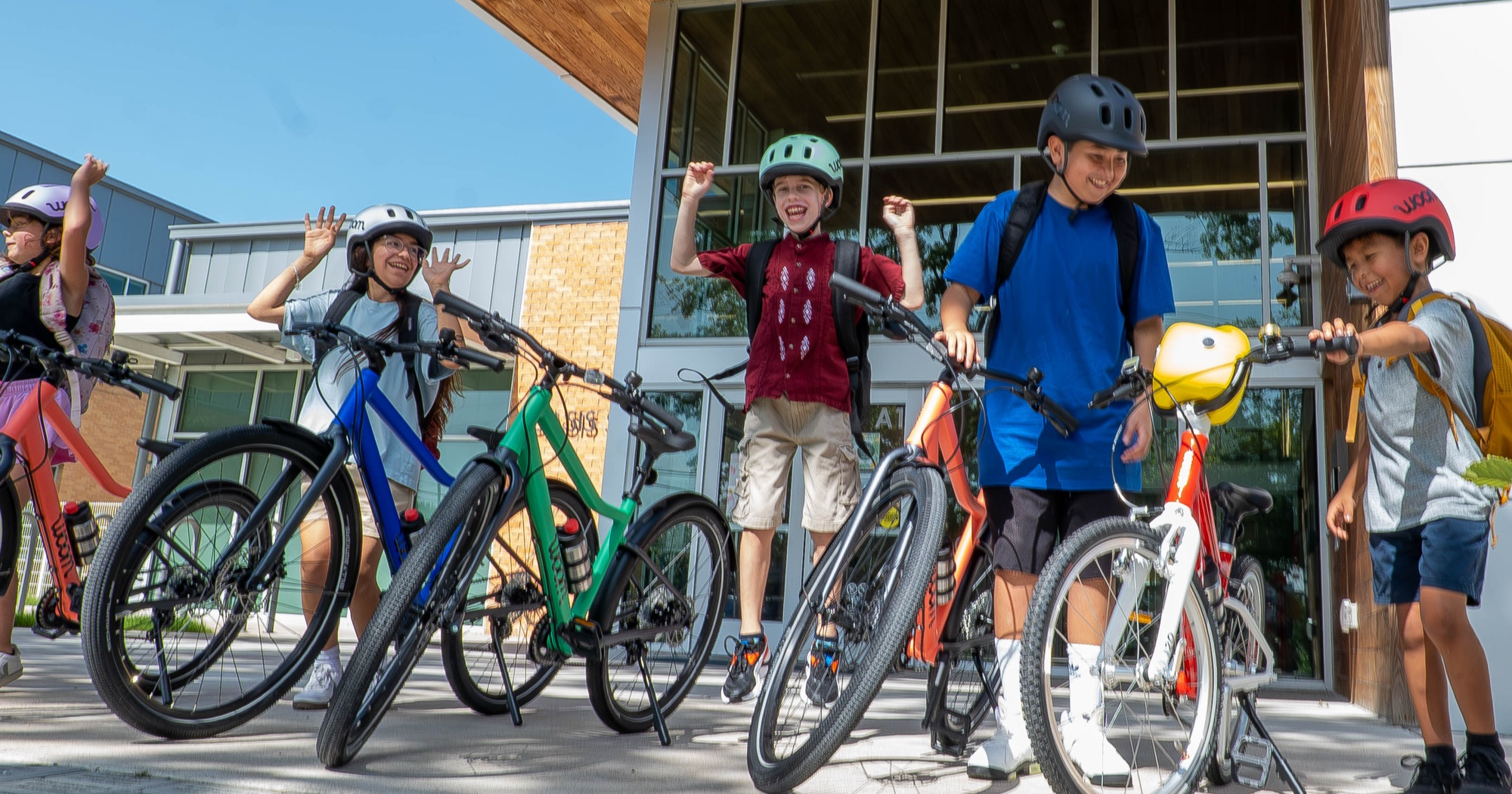 group of kids with woom EXPLORE and woom GO bikes to school while wearing woom READY Kids' Helmets and backpacks standing in front of a school