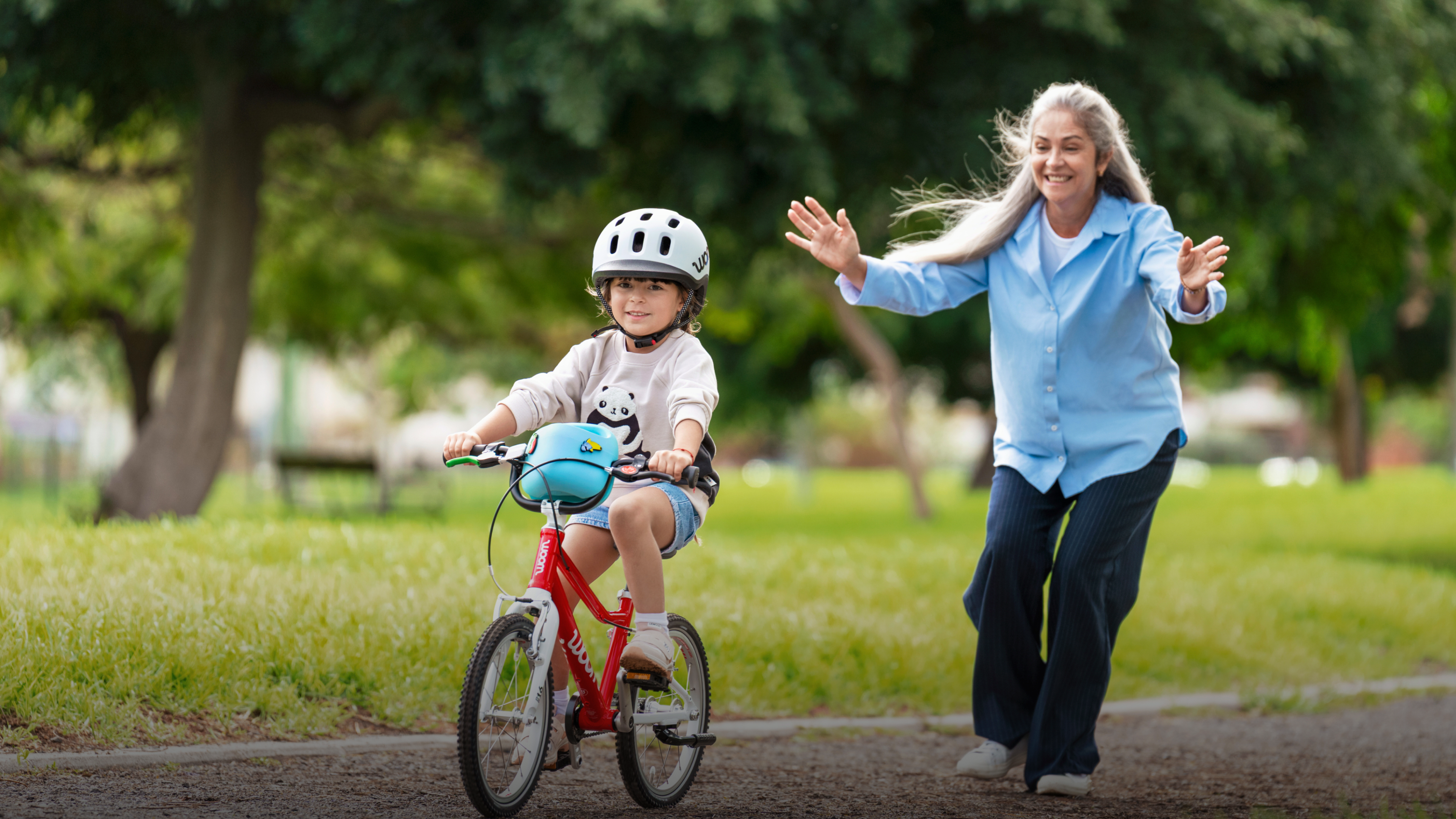 Child wearing a woom READY helmet riding a red woom GO bike with a woman watching in a park.