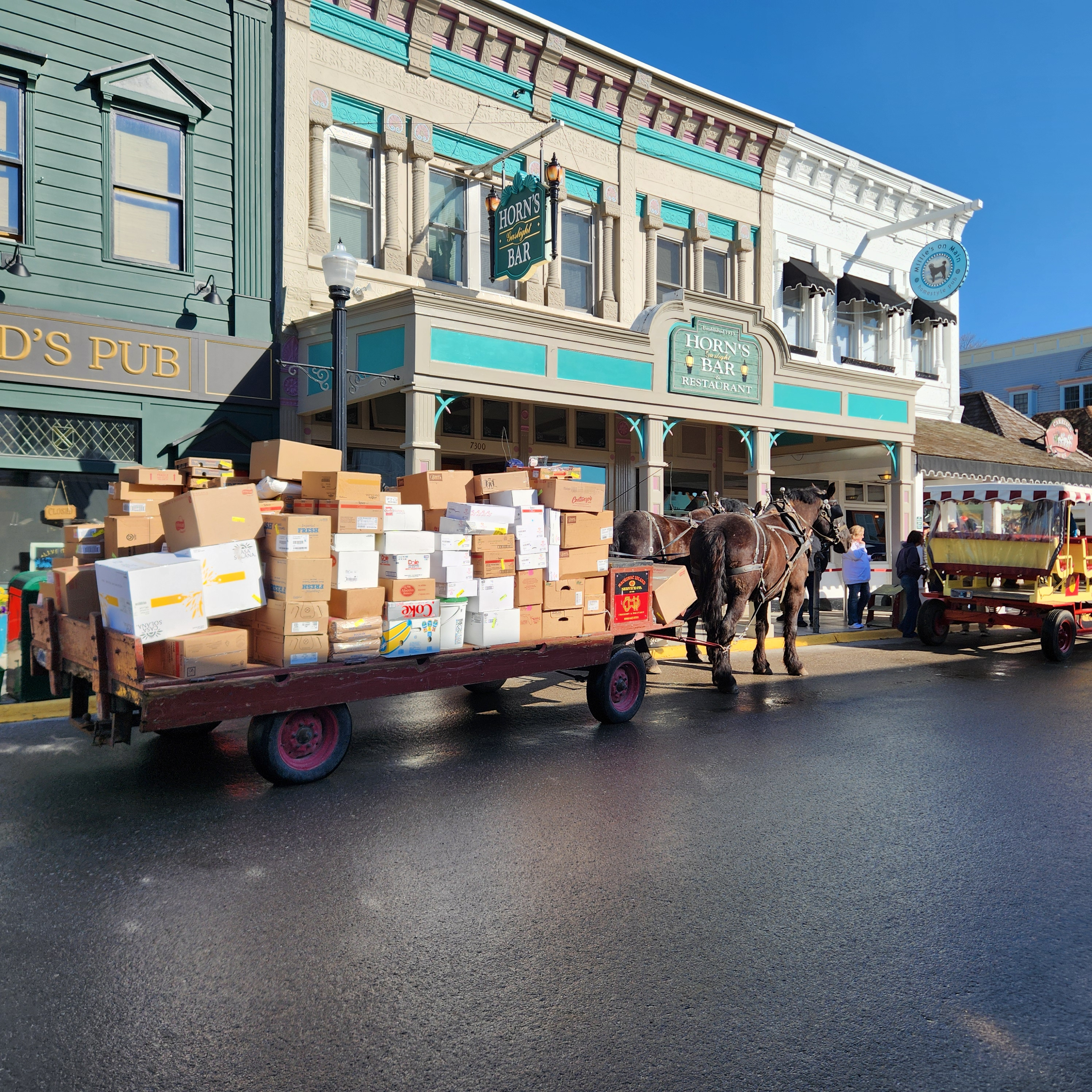  A horse-drawn trailer transporting numerous cardboard boxes is parked on the street in front of the teal and grey entrance to Horn’s Gaslight Bar & Restaurant on Mackinac Island.