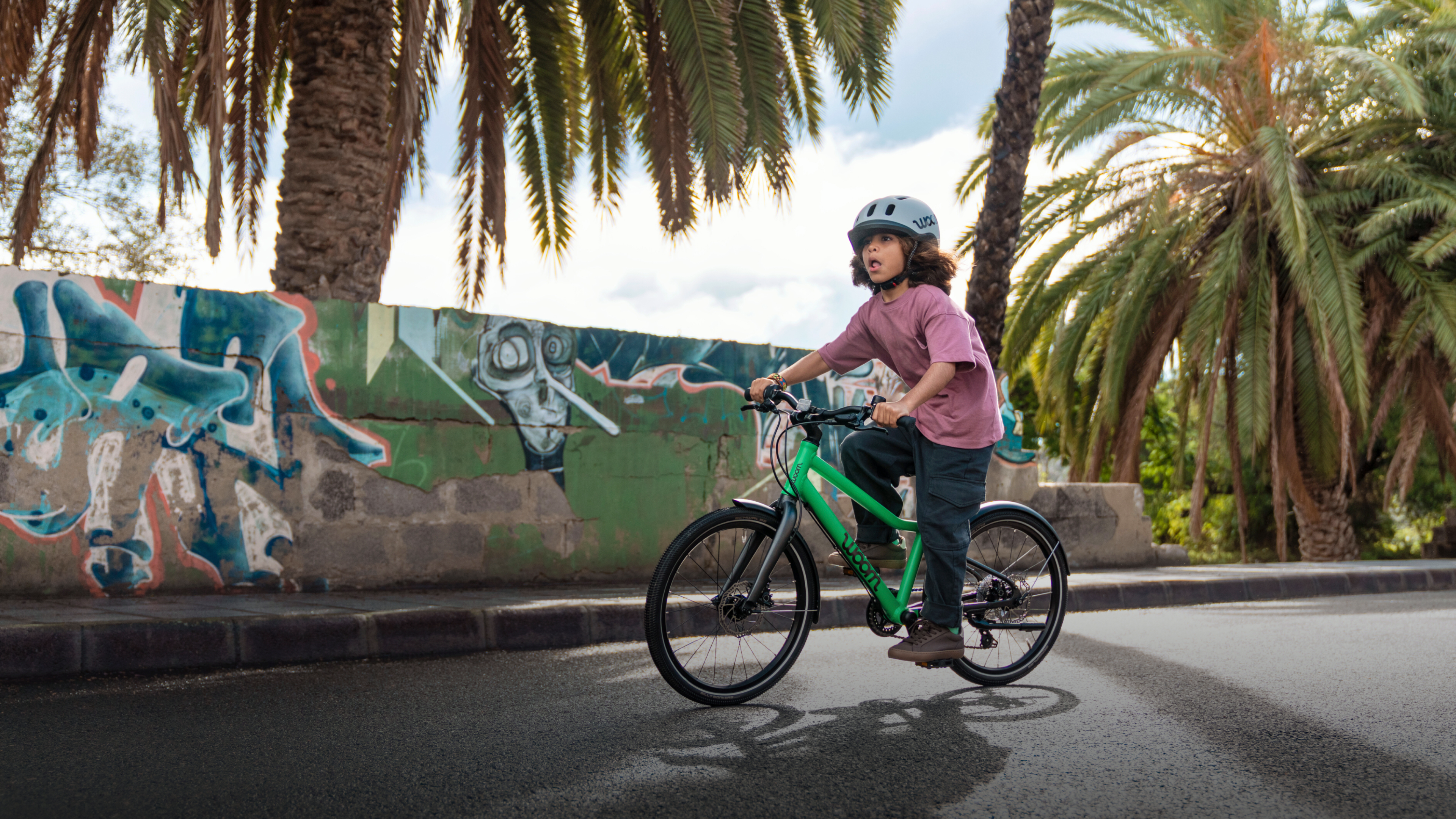 Child wearing a woom READY helmet riding a green woom EXPLORE bike past a graffiti wall.