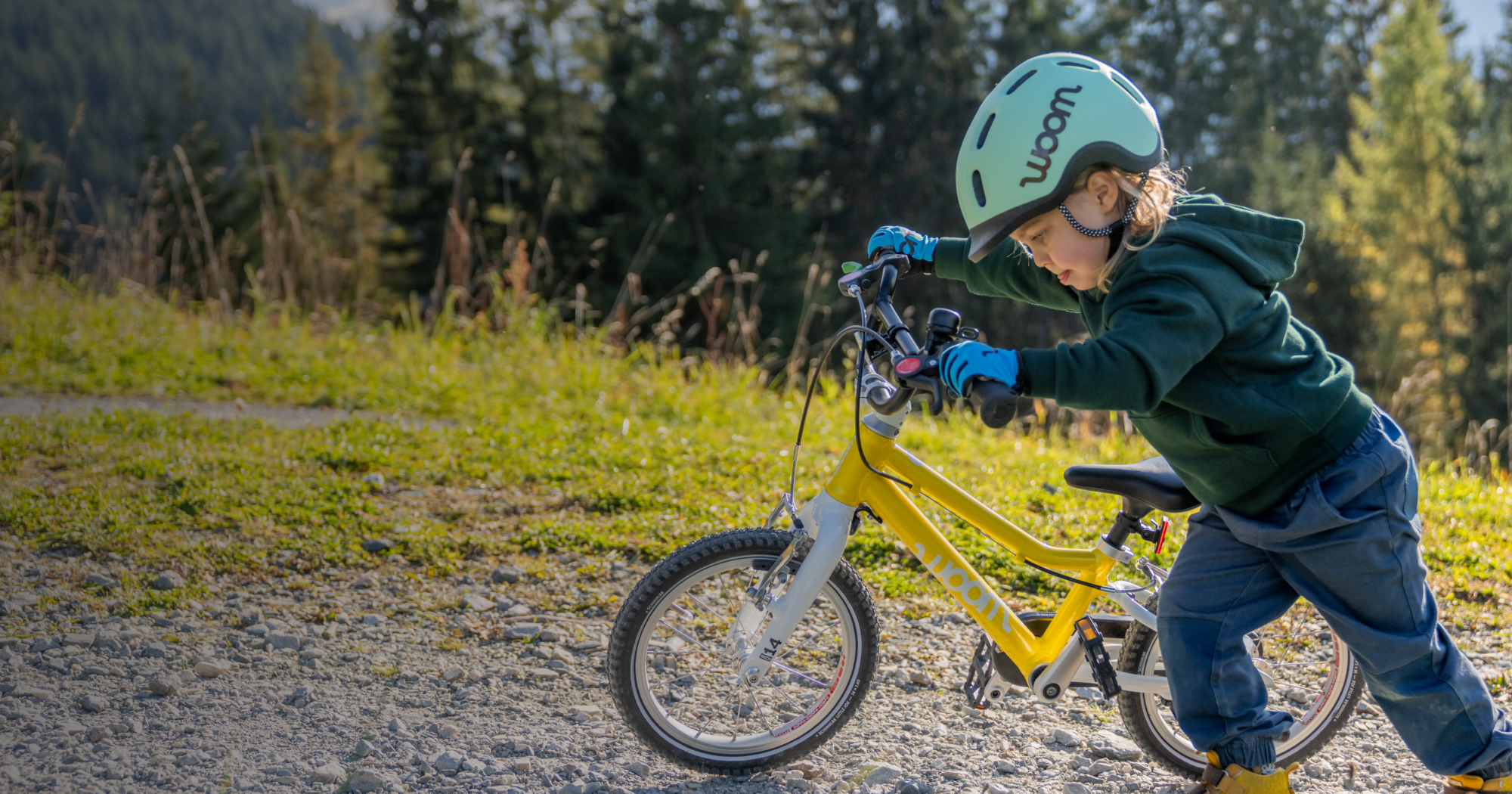 A young child in a mint READY Kids’ Helmet and sky blue TENS Bike Gloves focuses intensely on pushing his vibrant yellow woom GO 2 up a tree-lined incline.