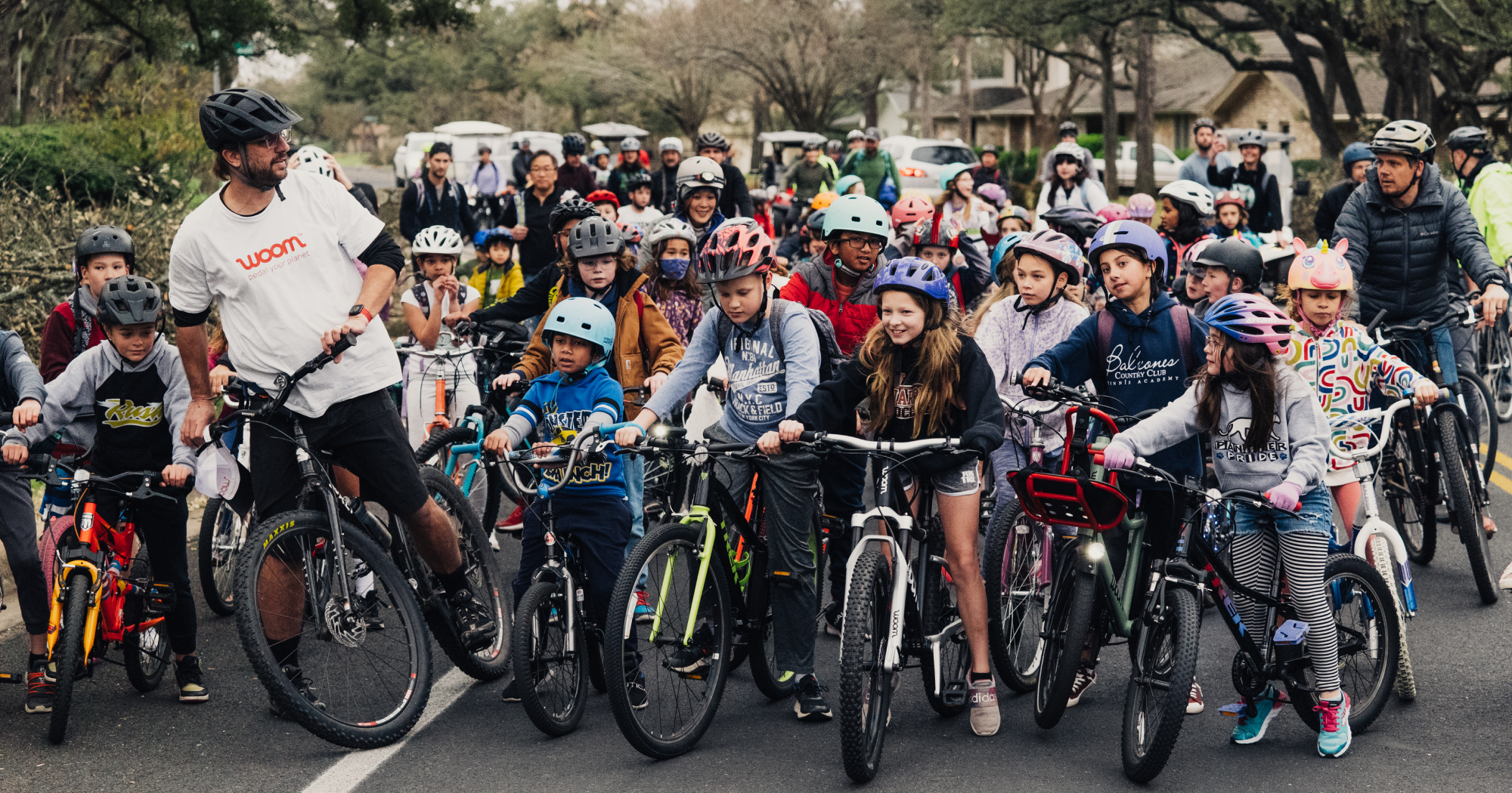 A crowd of kids on bikes lines up at the direction of their chaperone to start their Bike Bus ride on a neighborhood street.