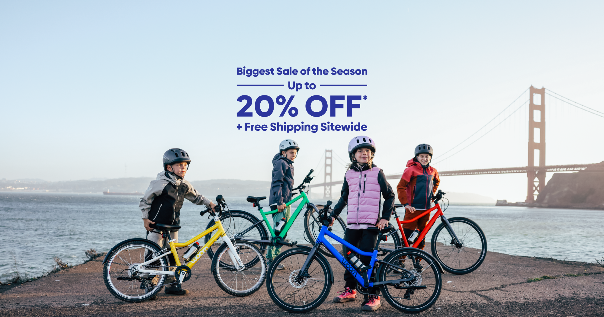 Kids pose with their colorful woom GO and EXPLORE bikes in front of the Golden Gate Bridge on a chilly day. Text over the image reads “Biggest Sale of the Season: Up to 20% Off Plus Free Shipping Sitewide”