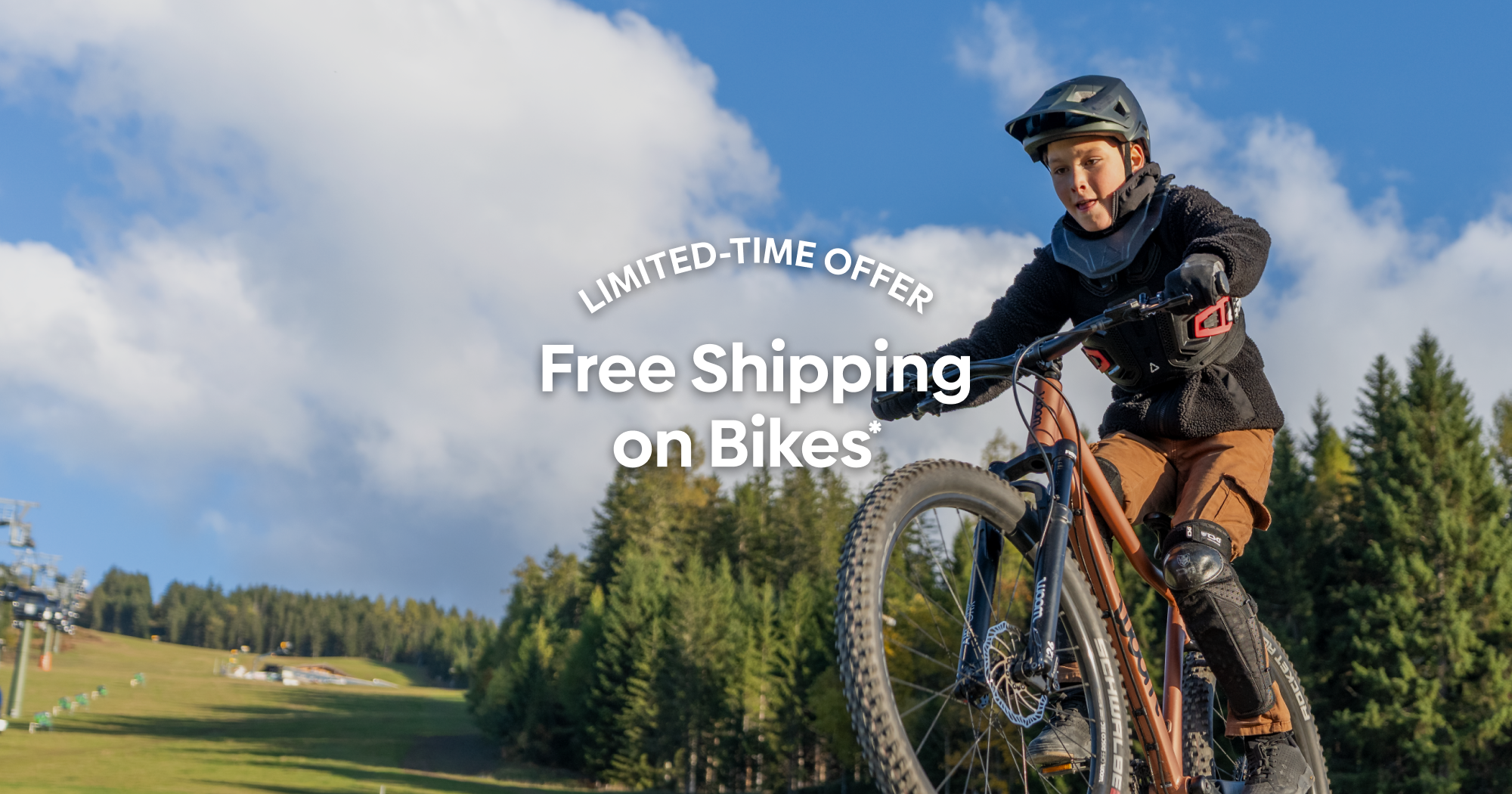 A boy in winter wear and a helmet fiercely concentrates while popping a wheelie on a terra coppa-colored woom OFF AIR mountain bike in a tree-lined lush green field.