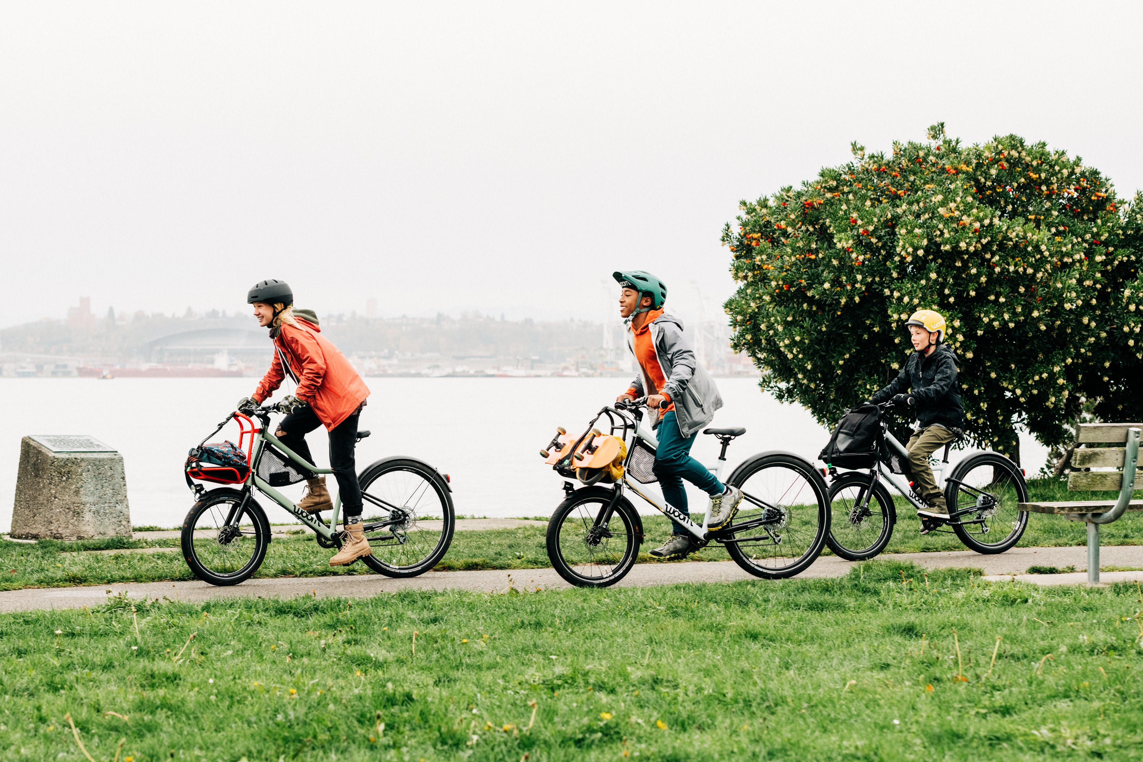 A group of kids riding a green and blue woom NOW city bike with a front rack beside a large river on a misty day.