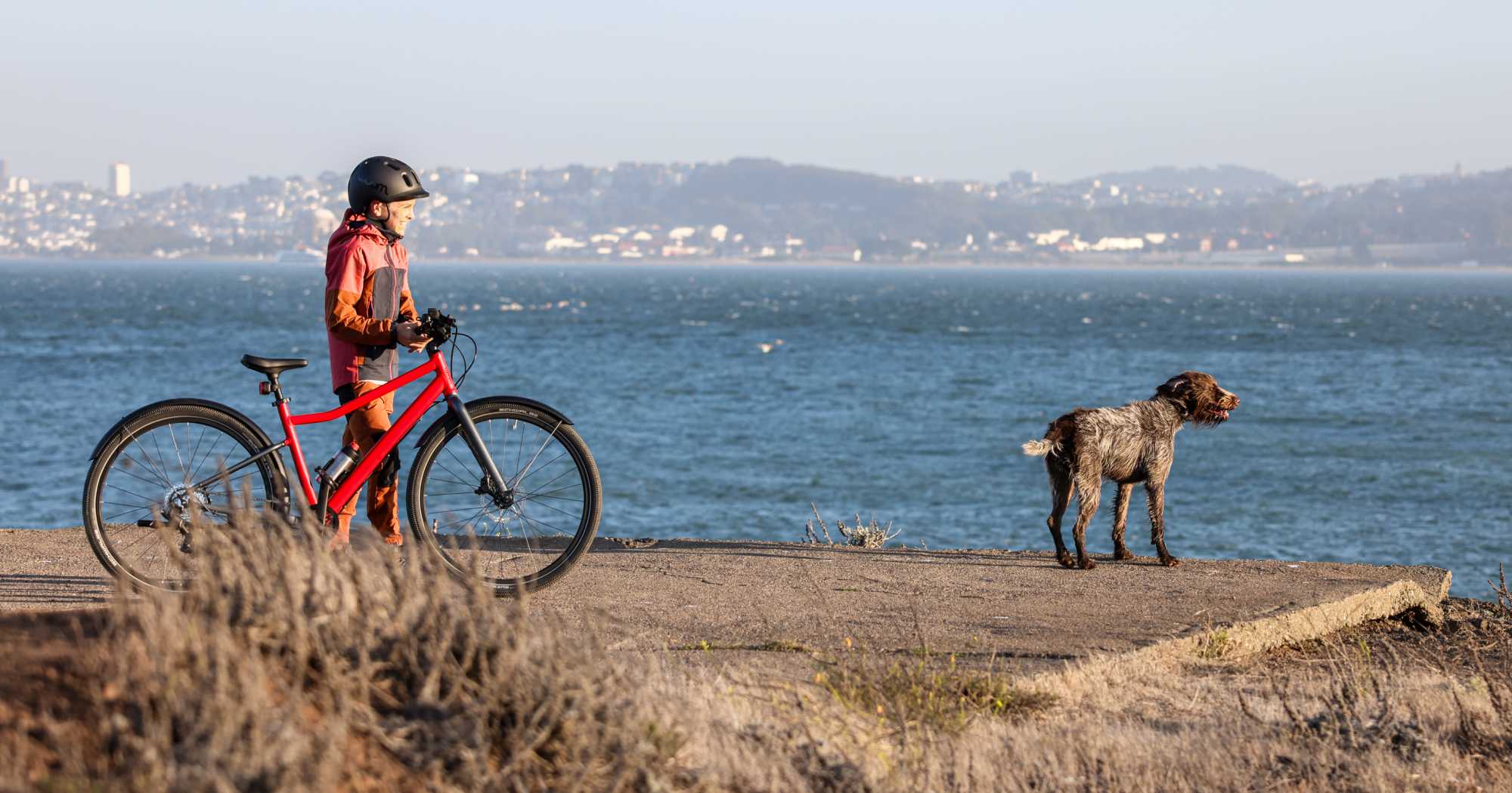 A boy wearing a READY Kids’ Helmet stands next to his woom red EXPLORE bike while looking out at the San Francisco Bay, accompanied by his dog