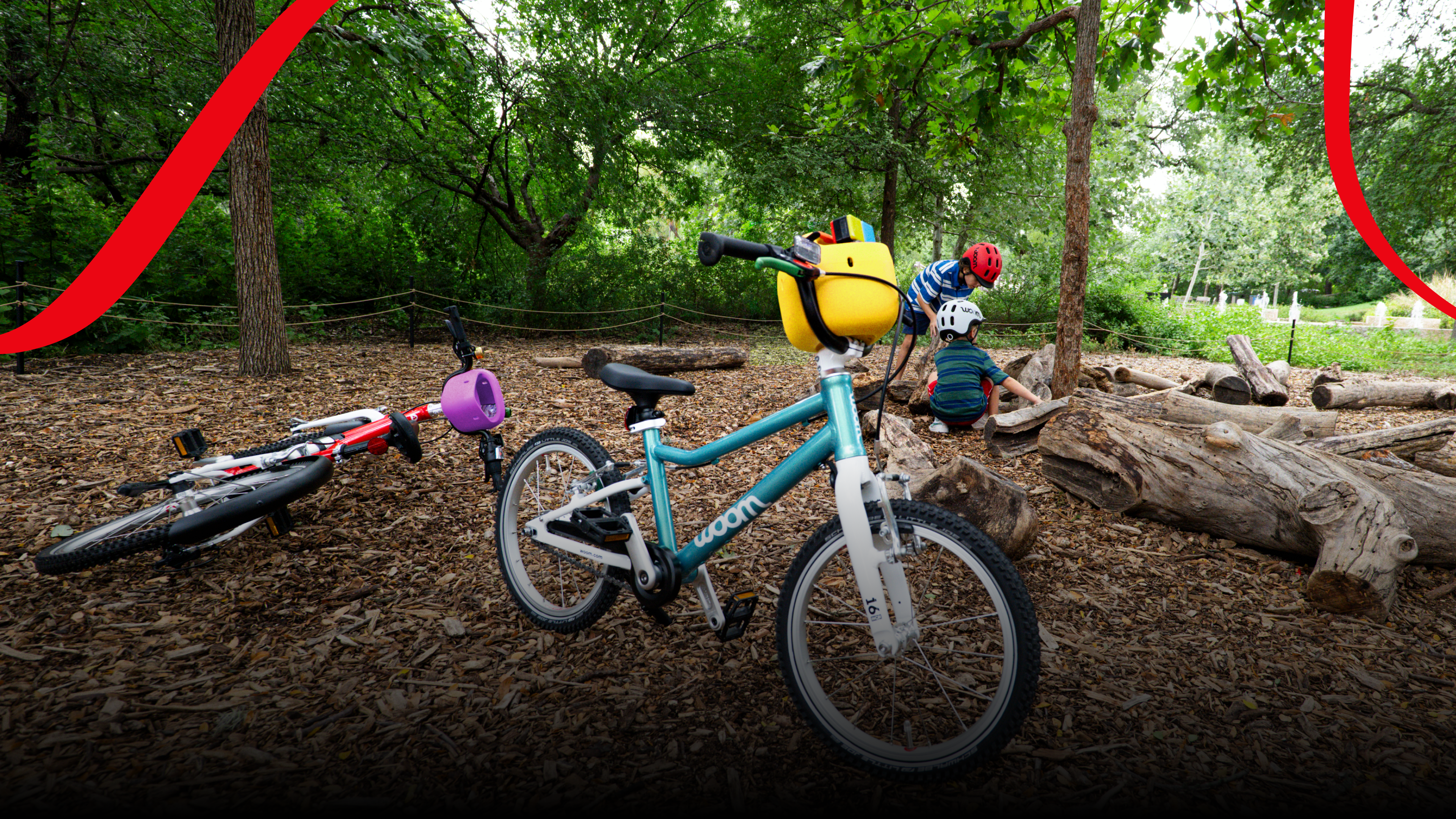 A woom GO in metallic turquoise with a yellow POP bicycle basket on the handlebars stands on a forest floor covered with wood chips, while two children wearing woom READY helmets play in the background.