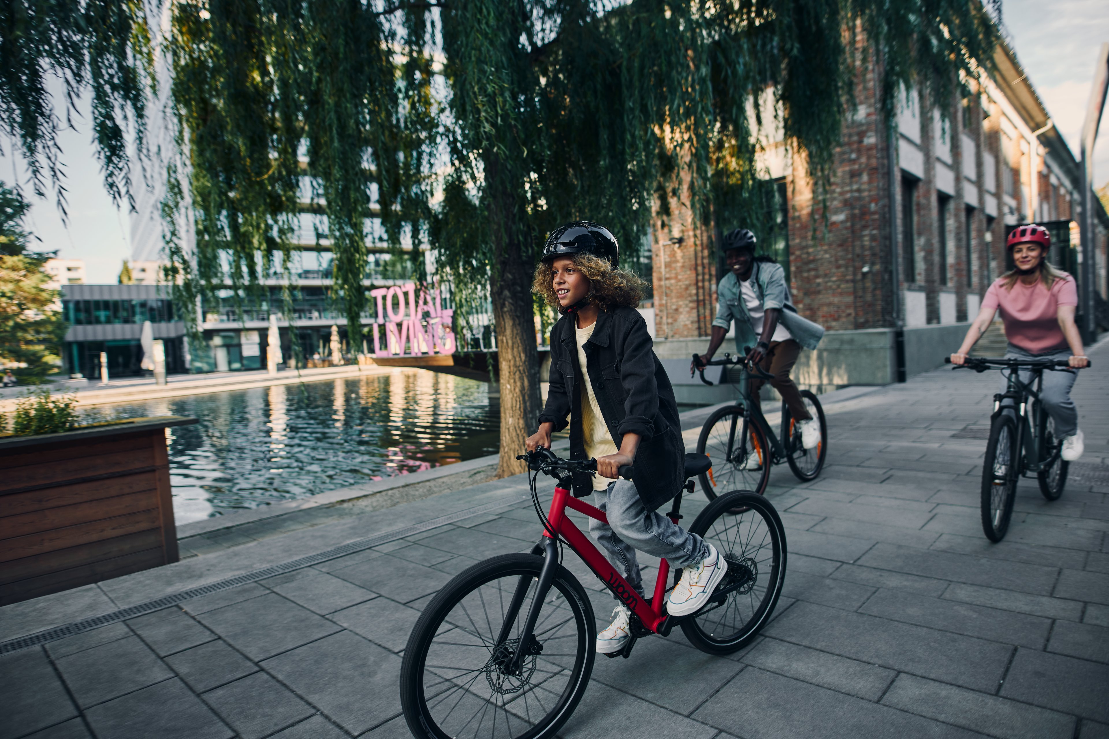 A child rides a red woom EXPLORE bike along the water, with two other people following behind on bicycles.
