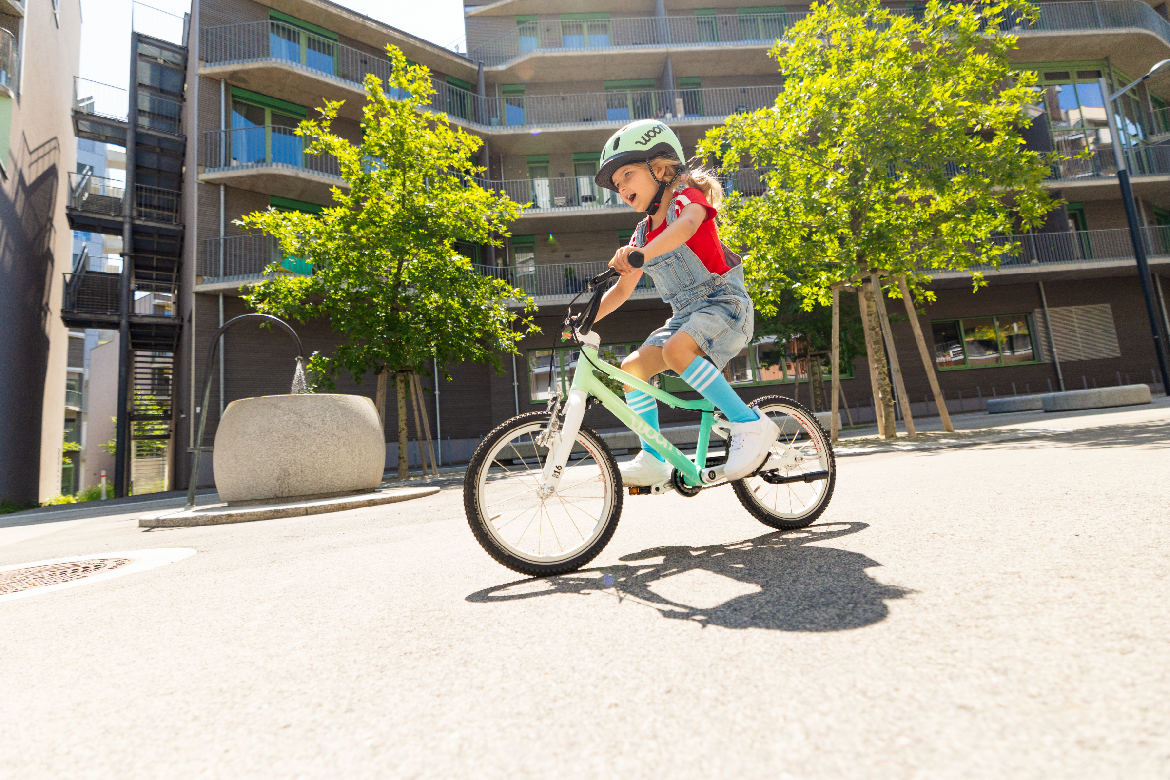 A girl wearing a mint-colored READY helmet rides past an apartment building on a spearmint crush woom GO.