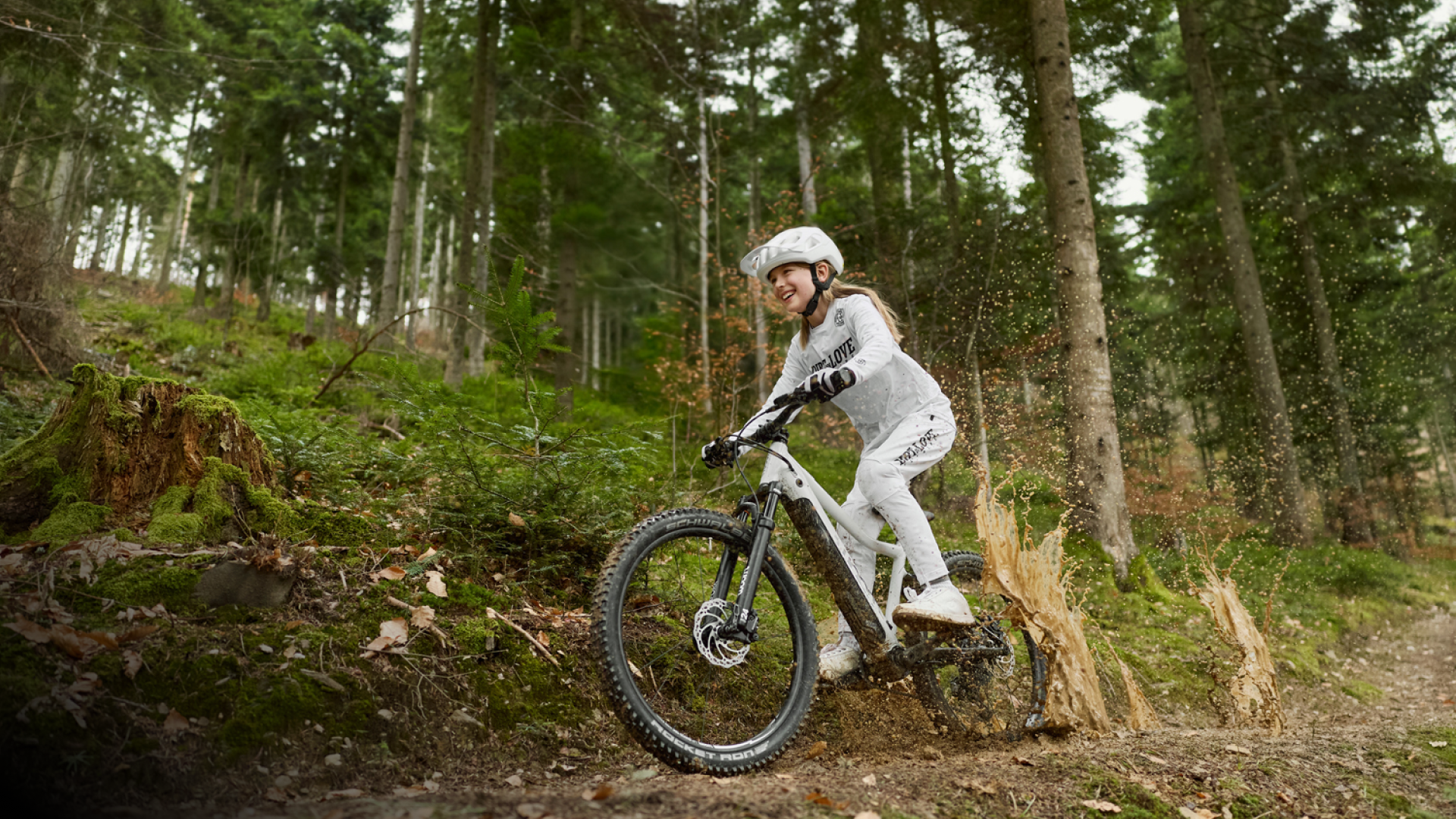 Lächelndes Mädchen auf dem E-Mountainbike woom UP in der Farbe cirrus cloud fährt über einen bewaldeten Trail.