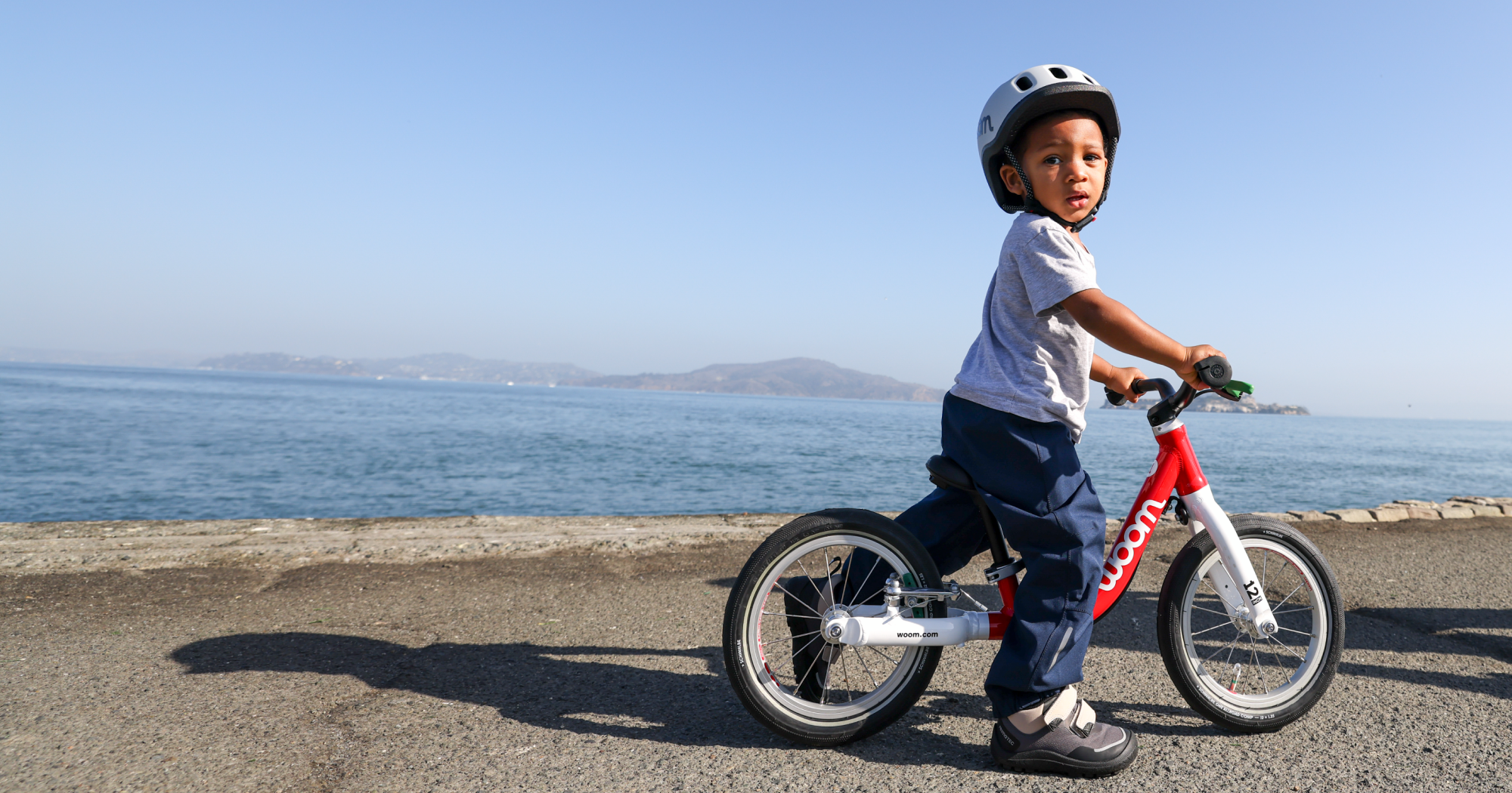 A boy wearing a woom grey READY helmet, riding a red woom GO bike by the San Francisco bay.