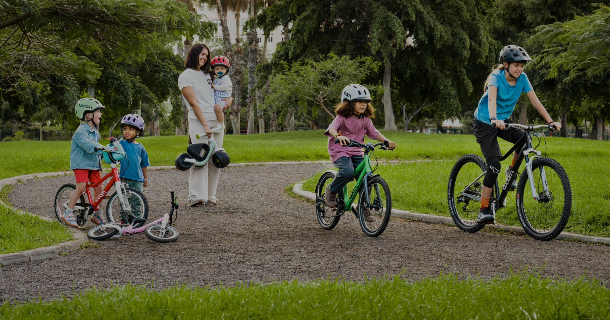 Multiple kids ride their woom GO, EXPLORE, and OFF bikes through a park while mom holds baby and a WOW balance bike.