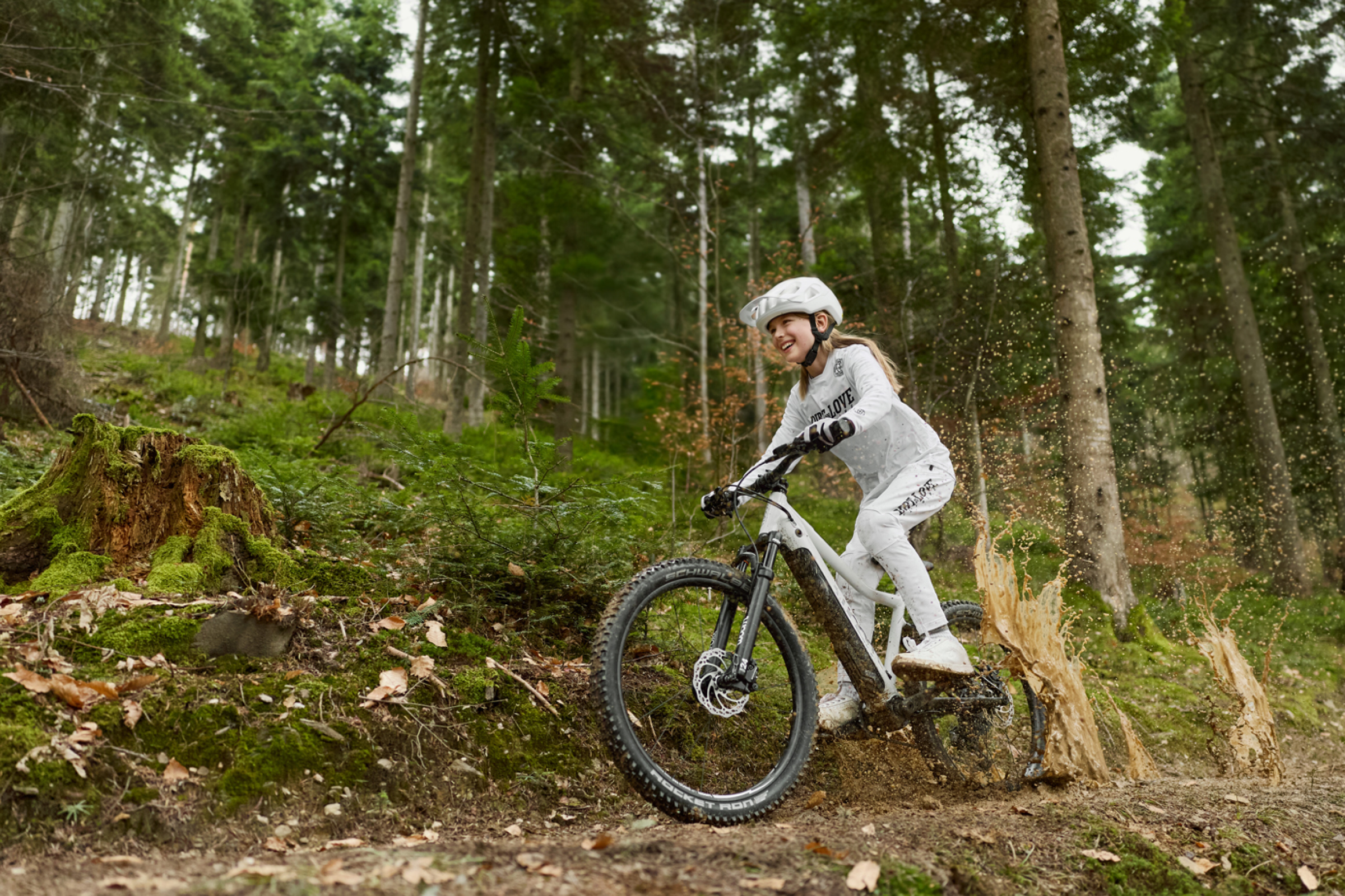 Ein lächelndes Mädchen fährt auf einem woom UP E-Mountainbike durch eine Pfütze im Wald.