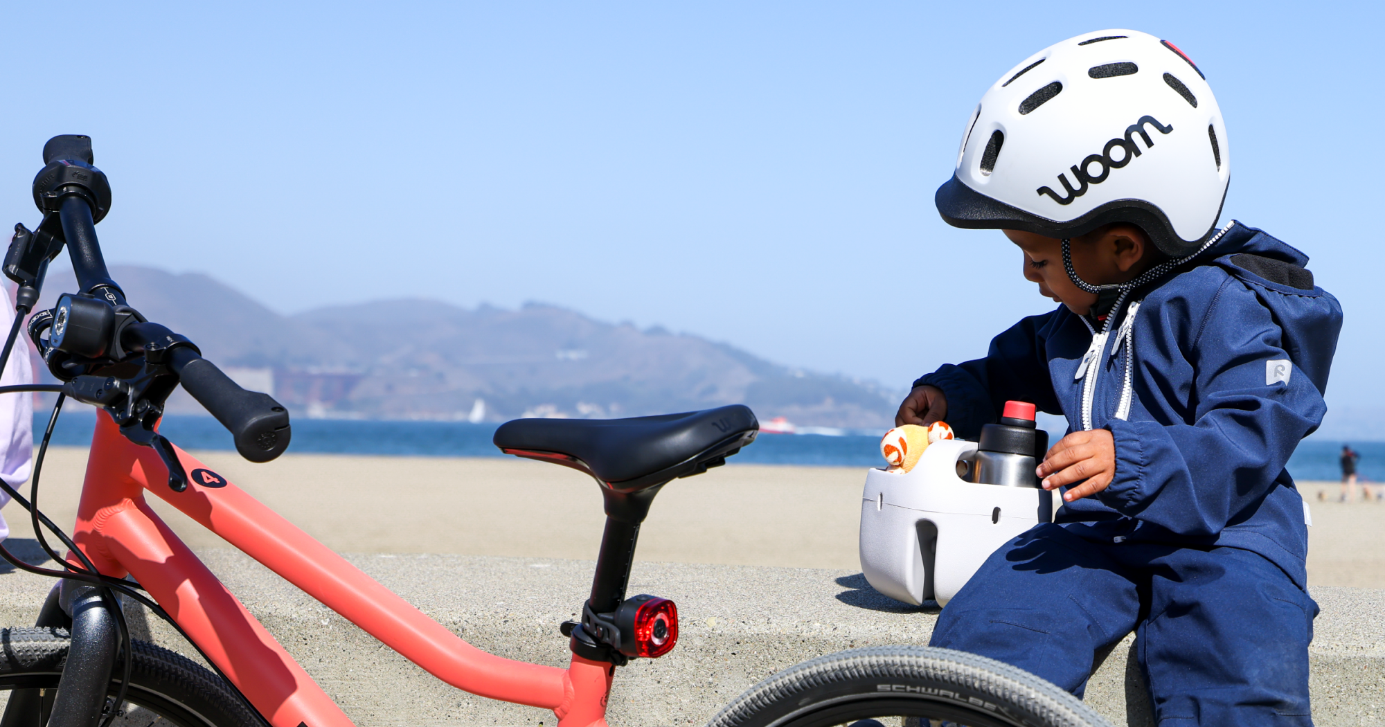 A young boy sits on a stone wall in front of San Francisco Bay looking at the contents of his POP Bike Basket next to his older sibling’s neon coral EXPLORE bike. He is wearing a READY Kid’s Helmet and his basket contains a stuffed animal and GLUG water bottle.
