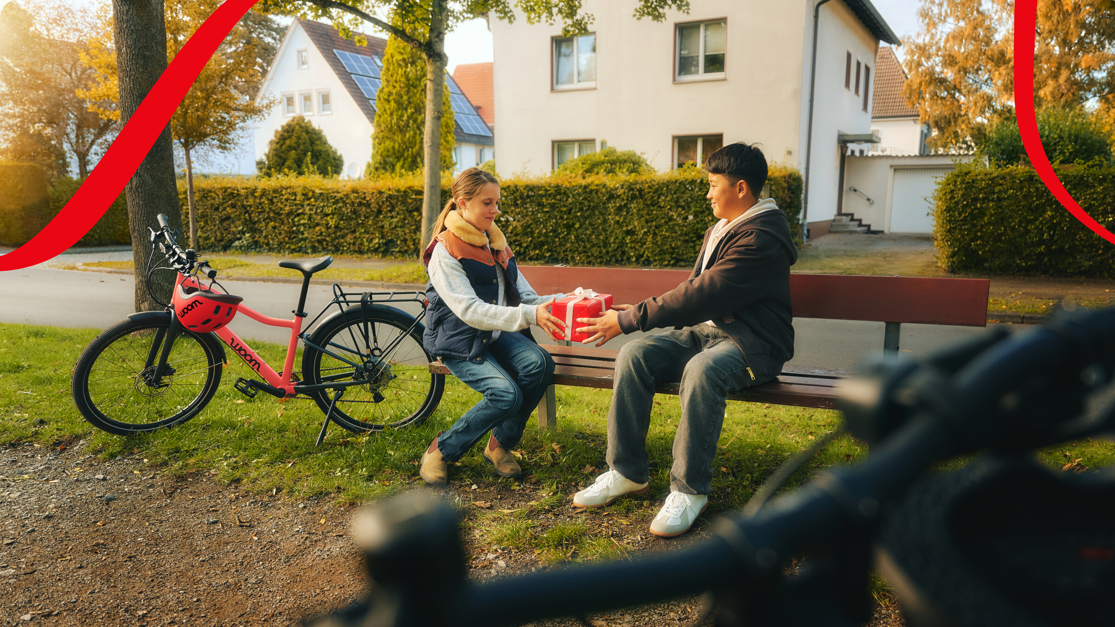 A boy and a girl are sitting on a park bench, and the boy is handing the girl a red-wrapped gift.