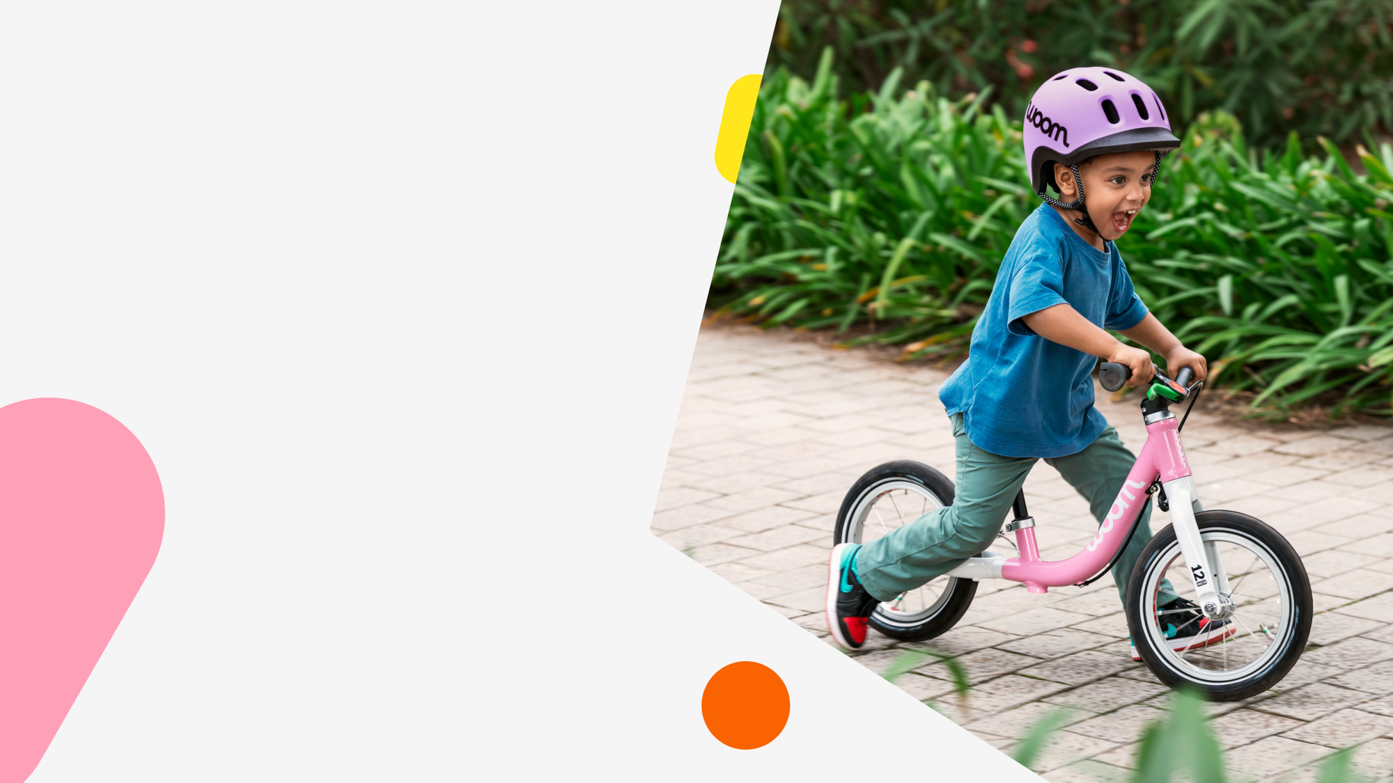 A young boy grins while riding a powder pink woom GO 1 down a brick path lined with bushes wearing a lilac READY Helmet