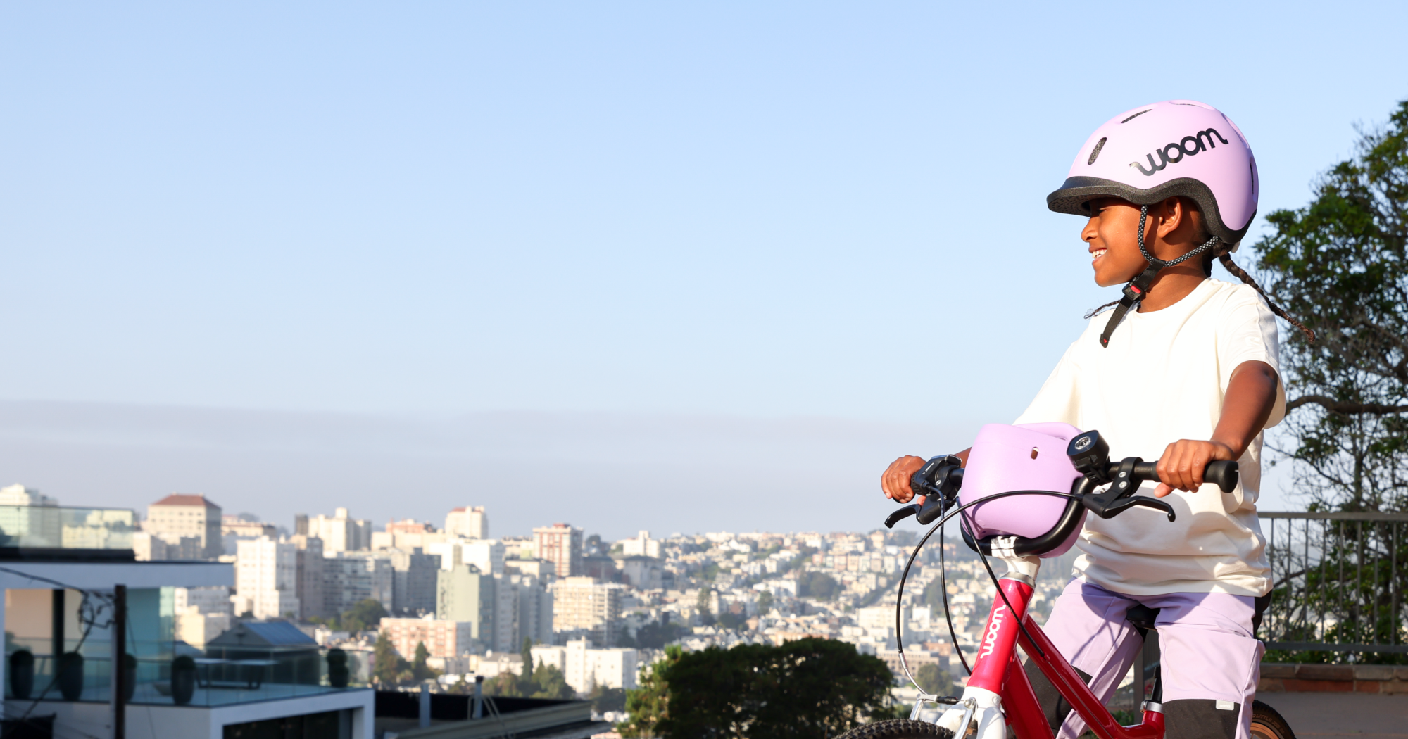 A young girl in braids and a lilac READY Kids’ Helmet sits atop her hot pink woom GO 4 bike, equipped with POP Bike Basket and BEAM Bike Light, overlooking a neighborhood from the top of a tall hill at golden hour.