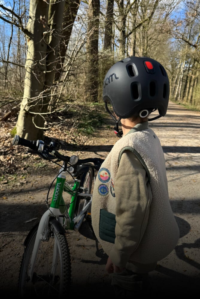 Et barn set bagfra med en sort READY hjelm på hovedet med integreret baglygte, woom cyklen er grøn.