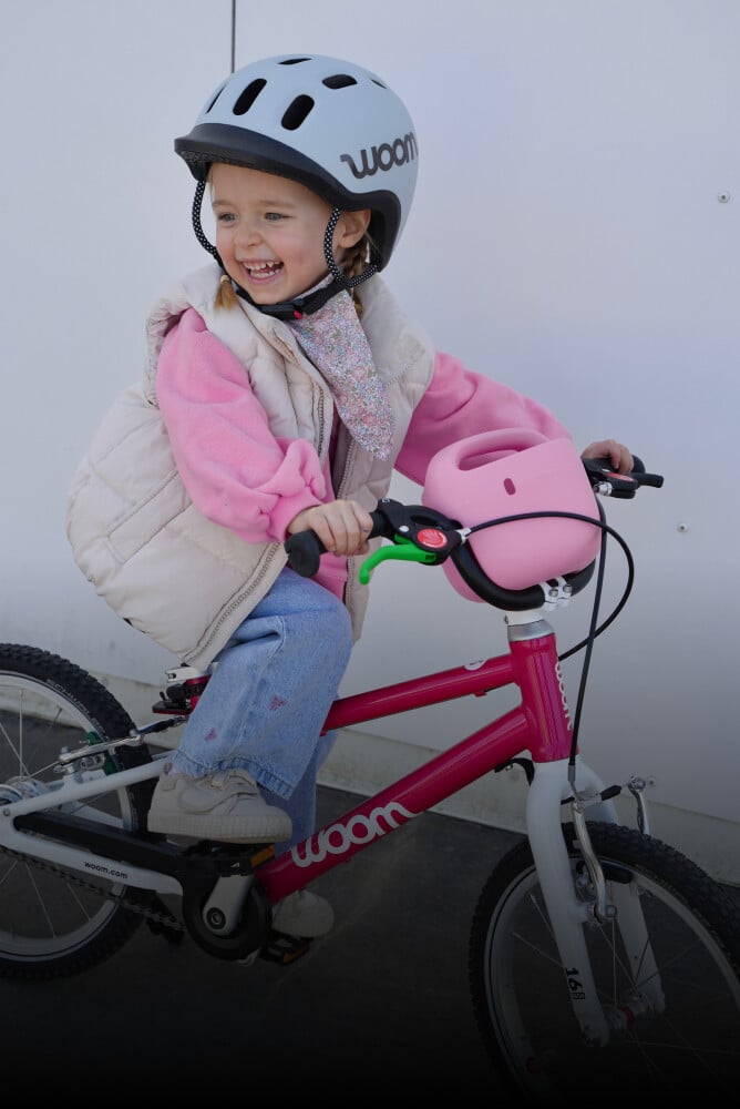En pige med en grå READY hjelm på hovedet sidder på en lyserød woom børnecykel med POP børnecykelkurv.