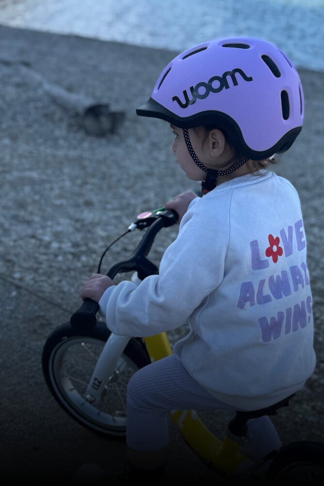 Et barn med en lilla READY cykelhjelm på hovedet sidder på en gul løbecykel fra woom.