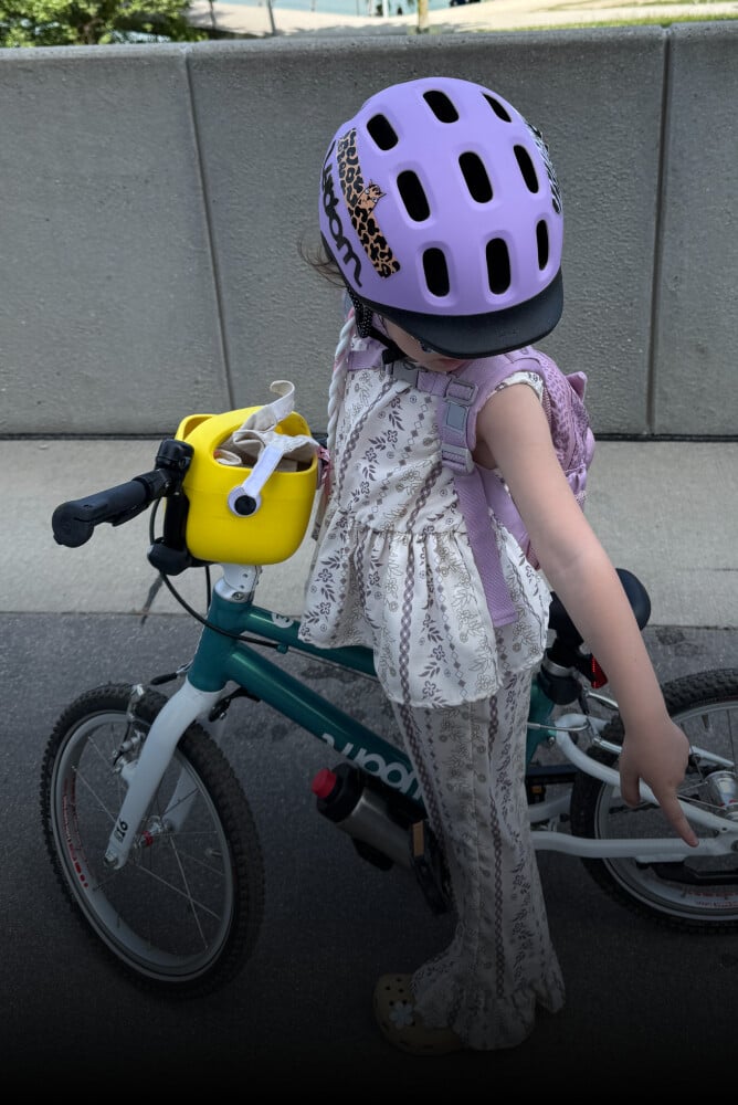 Barn med lilla READY cykelhjelm på hovedet står op på en grøn cykel fra woom med gul POP børnecykelkurv.