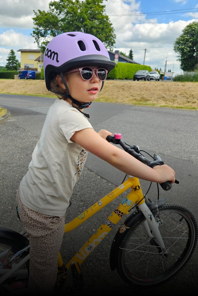 Et barn med lilla READY cykelhjelm på hovedet står op på en gul woom cykel med klistermærker på stellet.